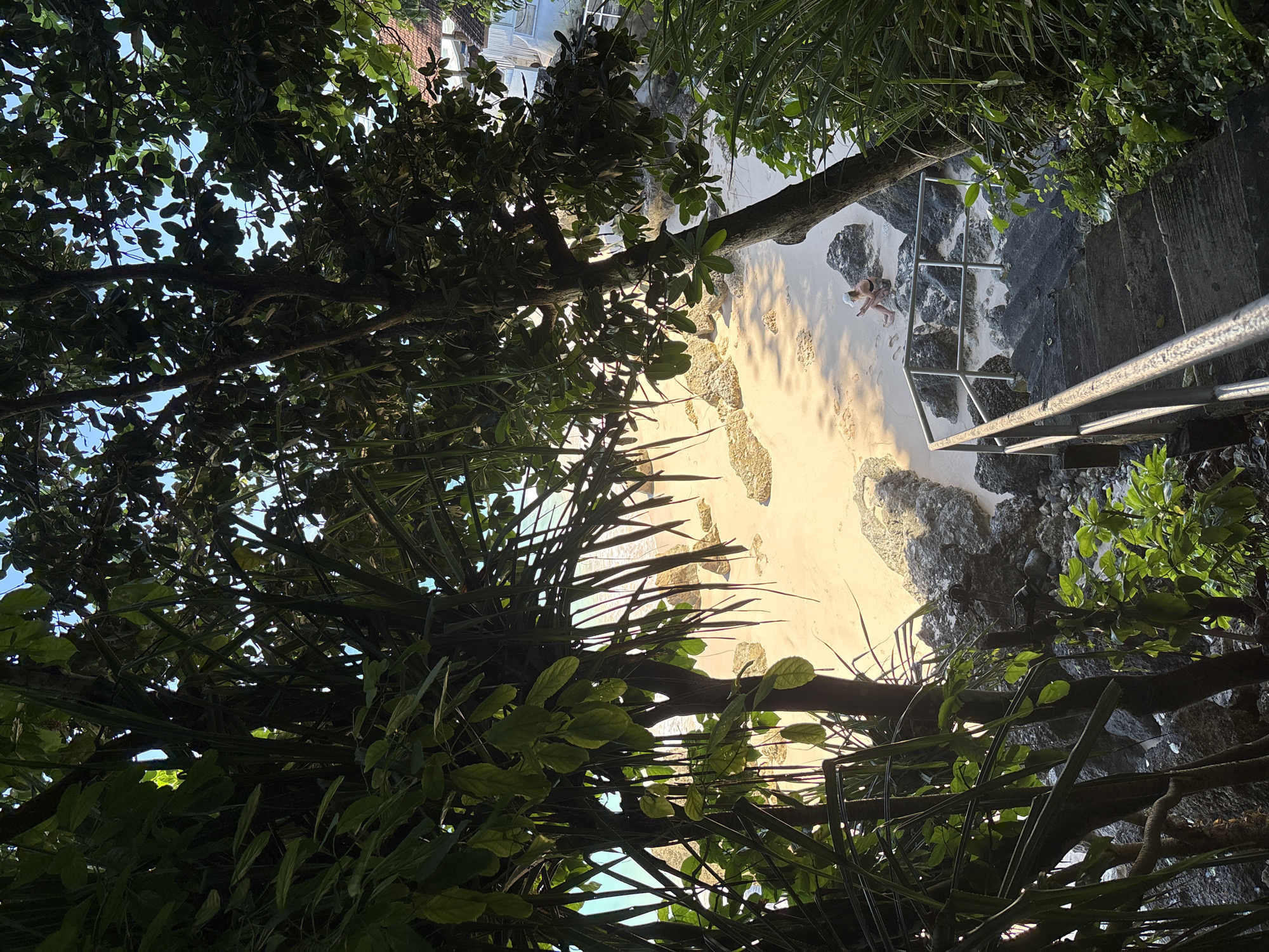 From an elevated vantage point amid dense foliage, the image captures a sunlit beach scene. A woman is present on the beach. A set of stairs with metal railings descends toward the beach. This scene is located in Kuta Selatan, Indonesia.
