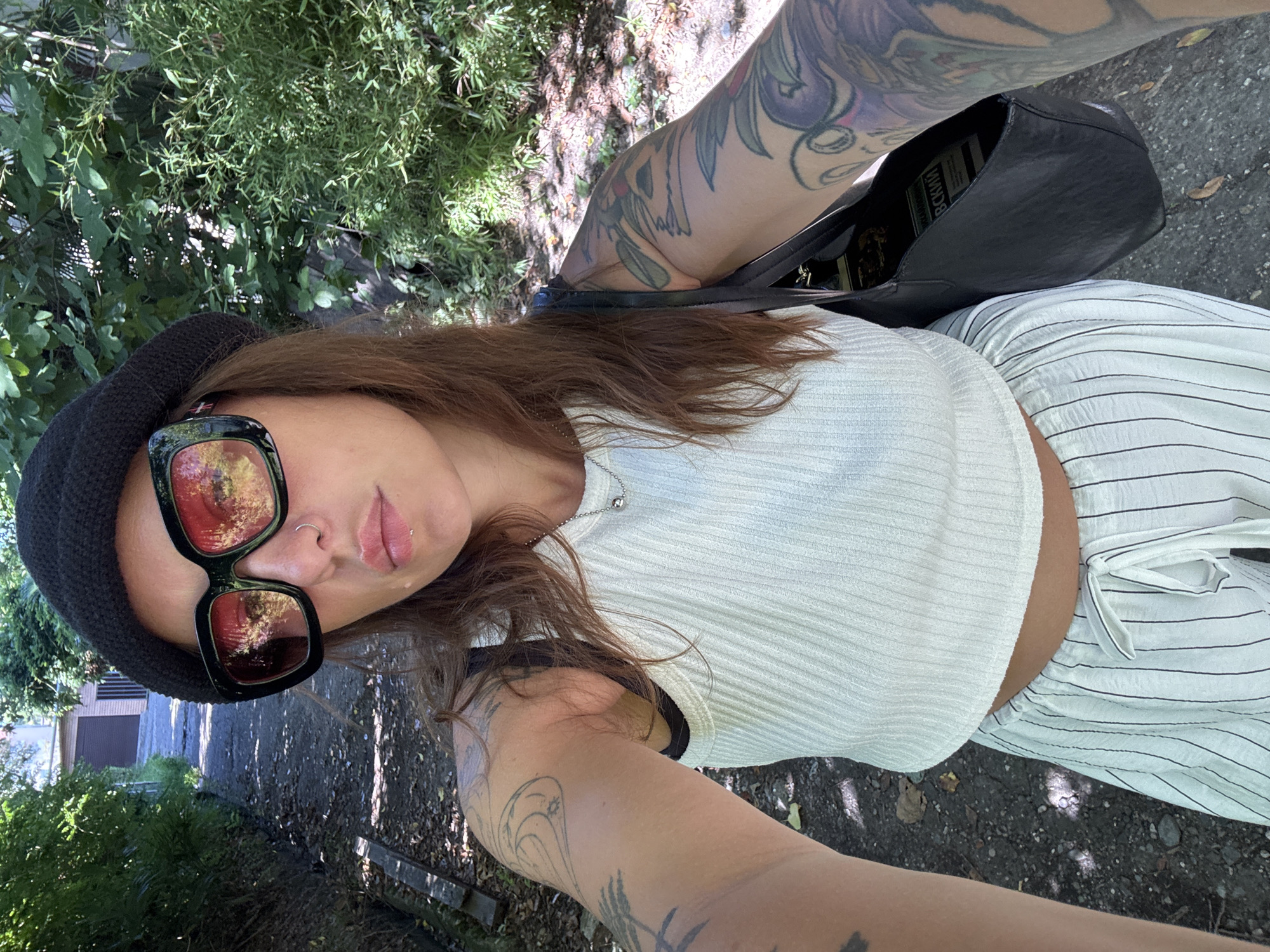 A young woman with long, brown hair and numerous tattoos on her arms is taking a selfie outdoors. She is wearing a white ribbed tank top, striped pants, a black beanie, and large, black-framed sunglasses with red lenses. A silver necklace with a small, spherical pendant is visible. She is also carrying a black shoulder bag. The background suggests an outdoor setting with a path leading uphill, bordered by lush green foliage and trees. A building with a dark roof is partially visible at the top of the path. The scene appears to be a sunny day, with dappled sunlight on the path and foliage. The location is identified as Sochi, Russia.