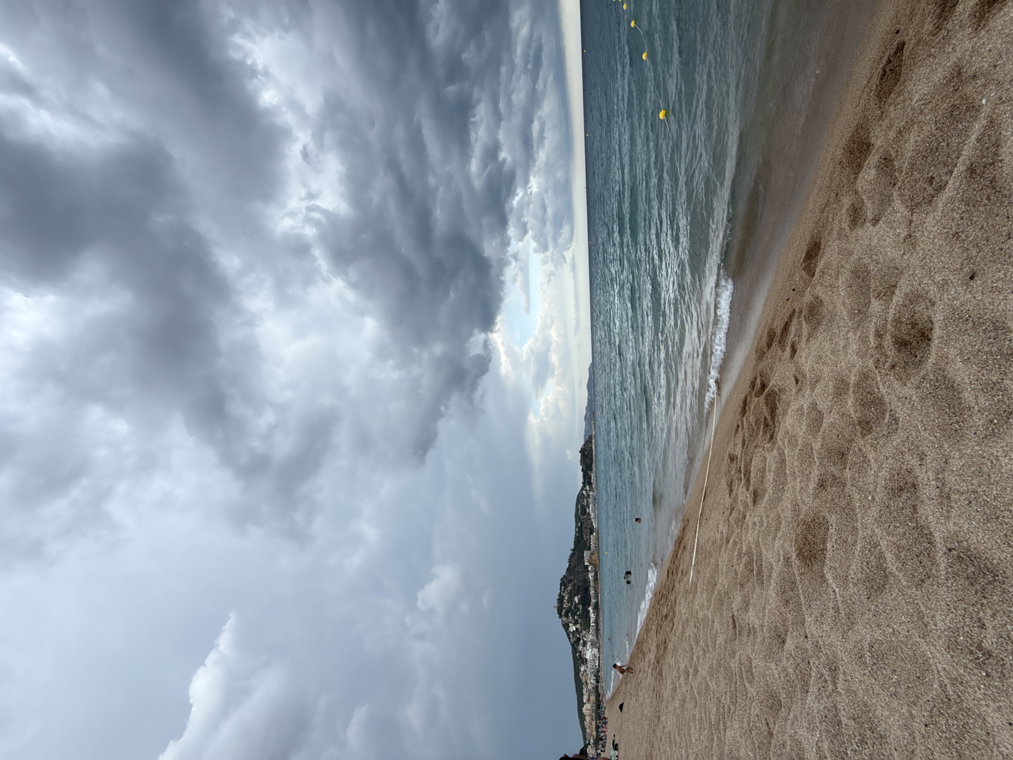 This is a photograph of a beach in Blanes, Spain. The foreground is dominated by a sandy beach with numerous footprints. The midground features the sea with gentle waves. Several yellow buoys are visible in the water, marking a swimming area. In the distance, a coastal town is nestled on a hillside, with buildings cascading down towards the sea. The sky is filled with dramatic, dark grey clouds, suggesting an approaching storm, though a small patch of lighter sky is visible towards the horizon. A few people are present on the beach and in the water.