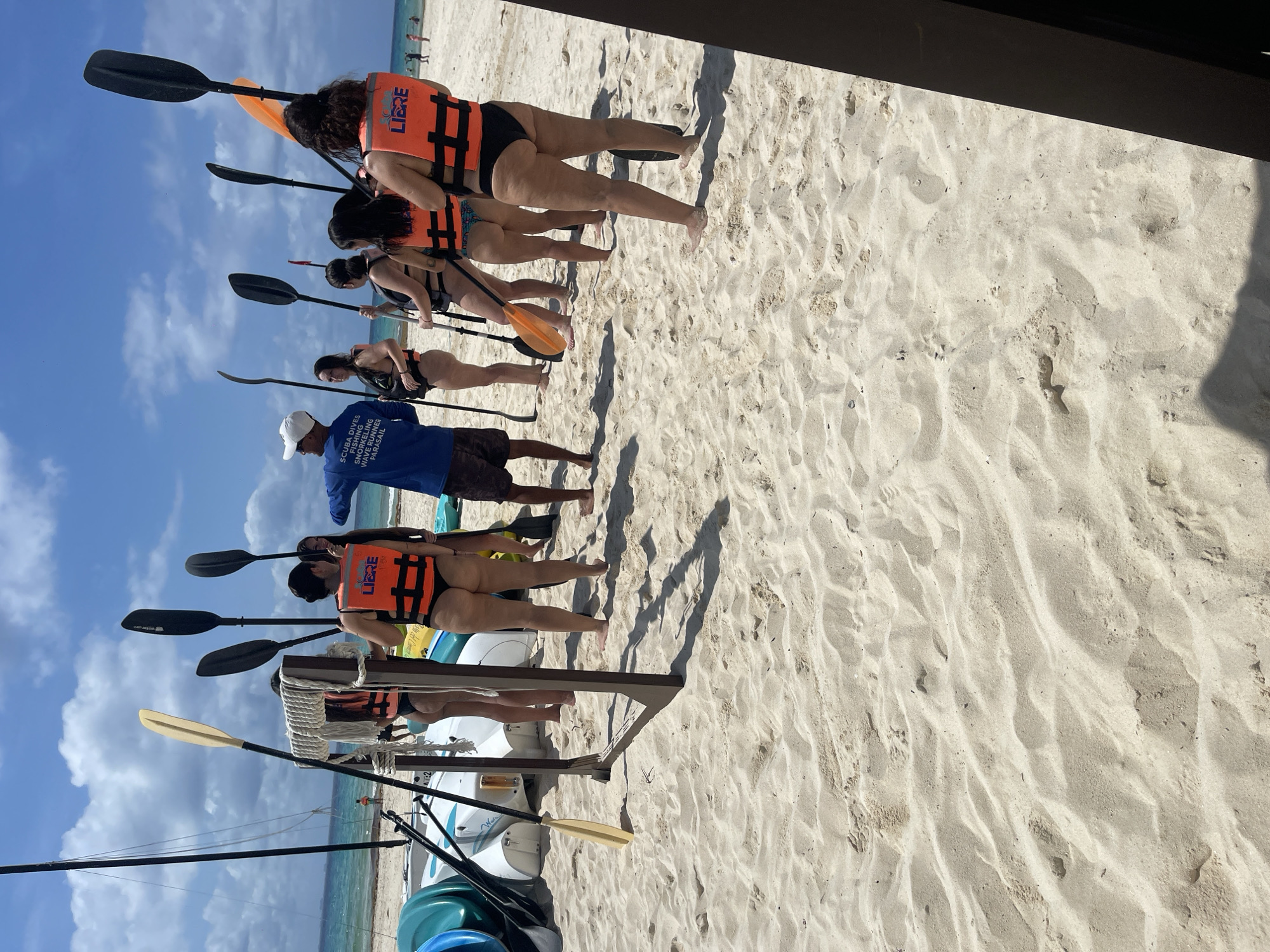 This image captures a scene on a sandy beach in Playa del Carmen, Mexico. Several women wearing life jackets and swimsuits are lined up holding paddles, presumably preparing for a kayaking or paddling activity. A man in a blue shirt and white cap appears to be instructing or guiding them. Kayaks and equipment are visible in the foreground, positioned on racks. The weather is clear and sunny, with a blue sky dotted with clouds. The sand is white and light. A partial view of the ocean is visible.