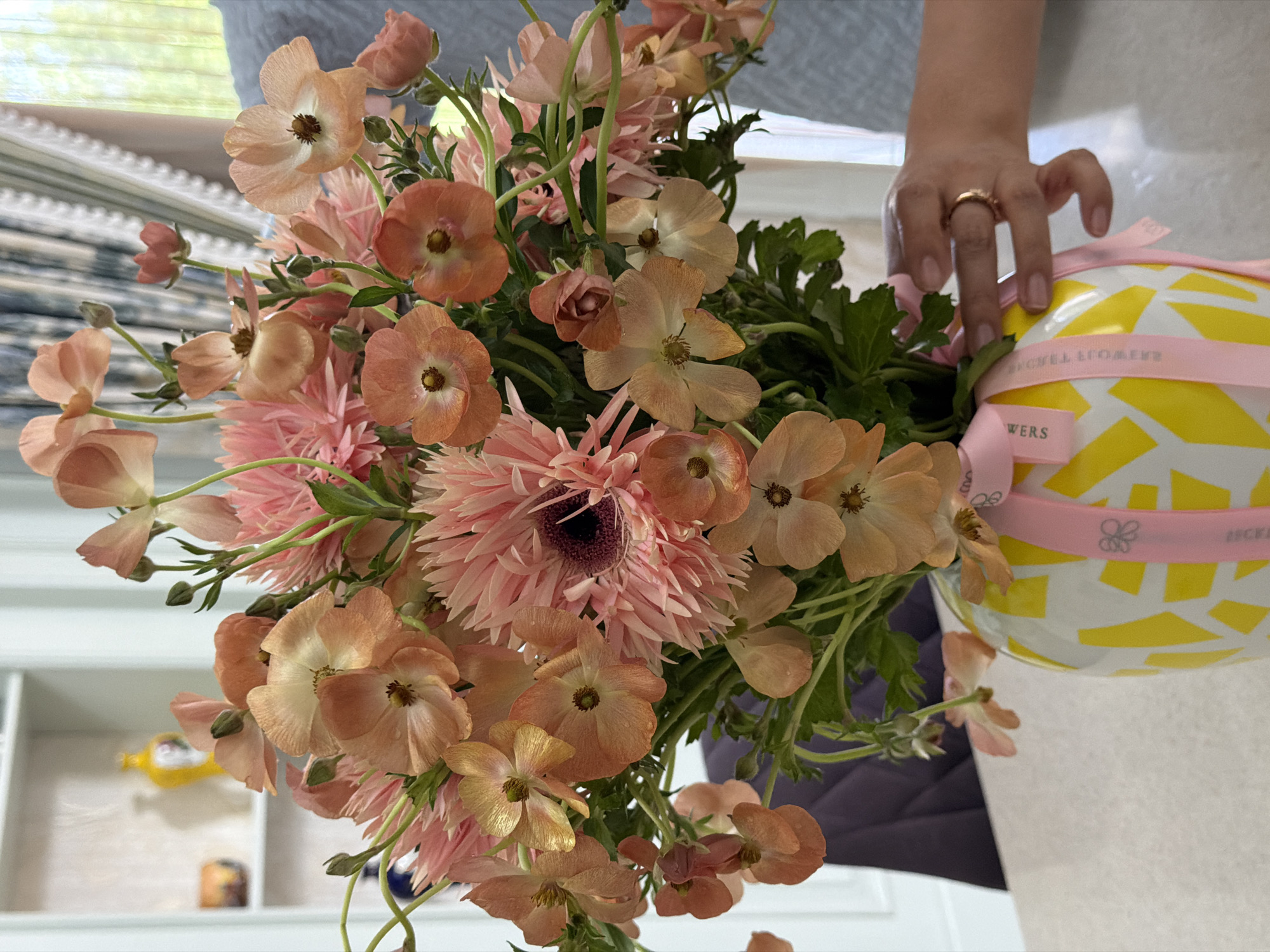 A close-up, eye-level shot showcases a vibrant bouquet of peach and pink flowers in a yellow and white patterned vase. The flowers, primarily ranunculus and gerbera daisies, are a mix of delicate peach petals and spiky pink blossoms. A pink ribbon with a floral pattern is tied around the neck of the vase, with visible text that appears to read "LOVELY FLOWERS." A person's hand, adorned with a gold ring, is gently holding the vase near the ribbon. The background is softly blurred, revealing hints of a light-colored shelf unit with decorative items, blue and white striped curtains, and a window allowing natural light to illuminate the scene. The overall setting suggests an indoor environment, likely a home or a flower shop, with the bright colors of the flowers and vase creating a cheerful and inviting atmosphere. The scene is clean and well-lit, with a focus on the floral arrangement.
