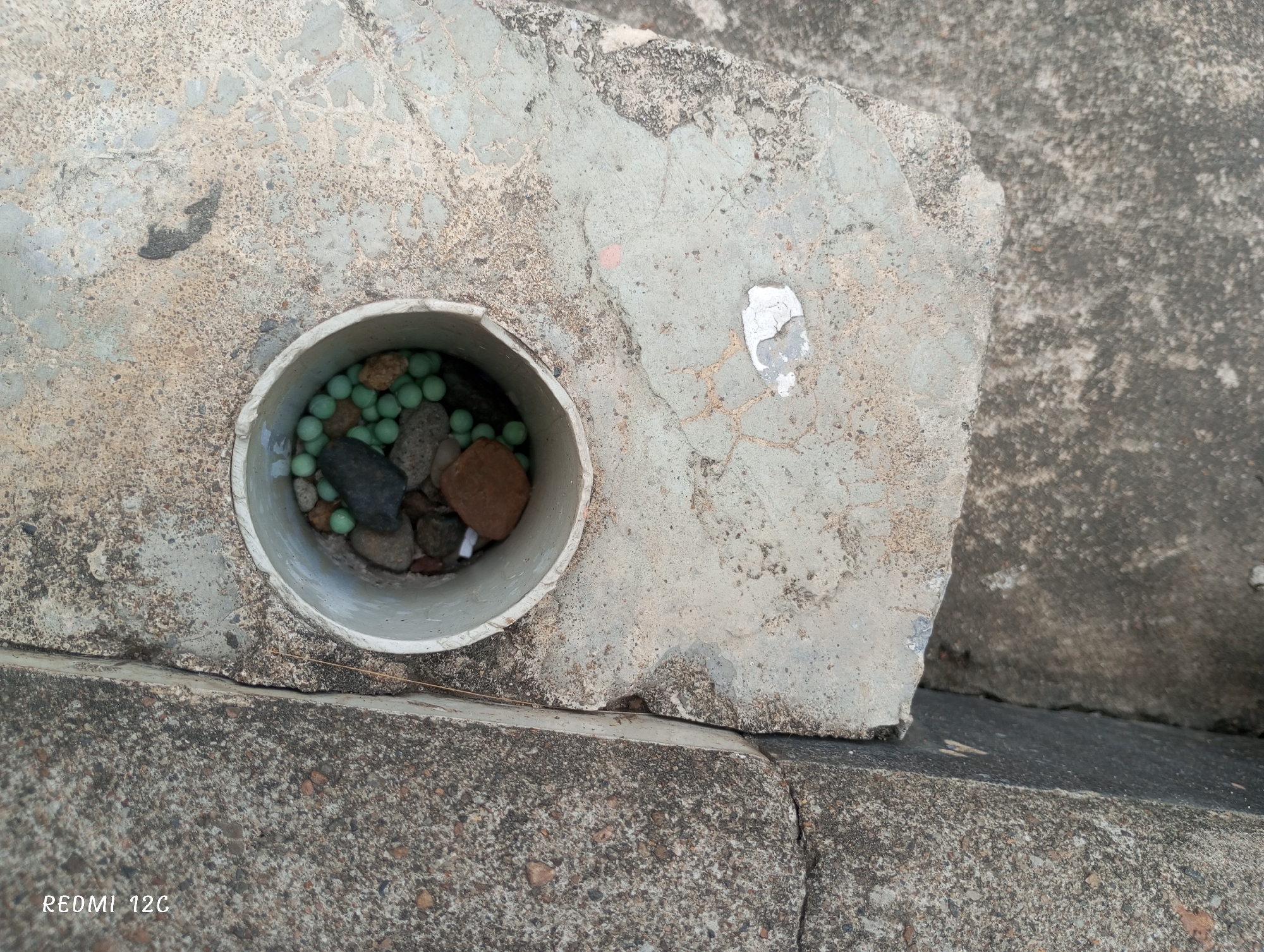 A close-up shows a segment of a concrete structure with a cylindrical pipe embedded in it. The pipe contains an assortment of small green spheres, rocks, and debris. The concrete surfaces are weathered and show signs of wear. The image was captured on a Redmi 12C phone. No people are visible. It is located in Lhokseumawe, Indonesia.