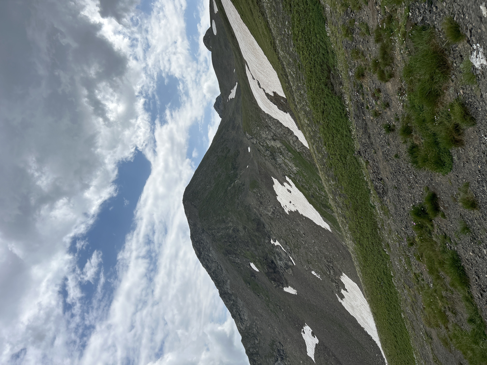 The image depicts a mountainous landscape under a partly cloudy sky. The foreground shows a rocky, gravelly slope with patches of green grass and small white wildflowers. Further up the slope, there are distinct bands of green vegetation interspersed with patches of snow, some of which appear to be melting. The mountain slopes are steep and rocky, with a prominent peak dominating the left side of the frame. In the distance, other mountain peaks are visible, also showing signs of snow. The sky is filled with a mix of white and gray clouds, with patches of blue sky visible. The scene appears to be a natural, undeveloped area in a mountainous region, likely Lunnaya Polyana, Russia, given the context. No people or man-made objects are visible in the image.