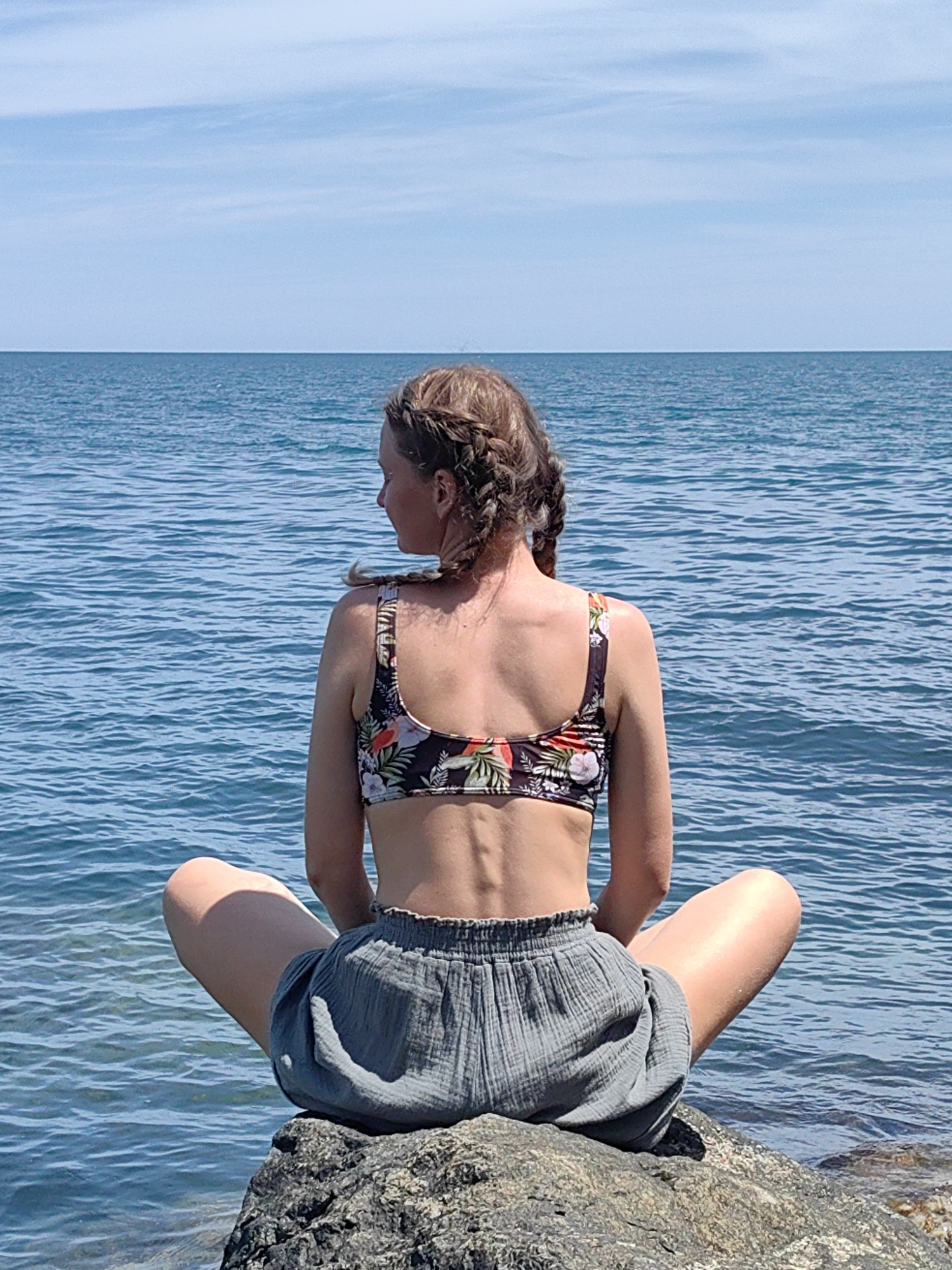 A woman with braided blonde hair sits in a cross-legged position on a rock by the sea. She is viewed from behind, facing the water. She wears a floral print bikini top and grey, crinkled shorts. The sea is a vibrant blue with ripples, and the sky above is a clear, pale blue with thin white clouds. The sun is shining, suggesting it is daytime, and the weather appears warm and pleasant. The overall scene evokes a sense of peace and relaxation, with the woman appearing contemplative as she gazes out at the ocean. There is no visible text in the image.