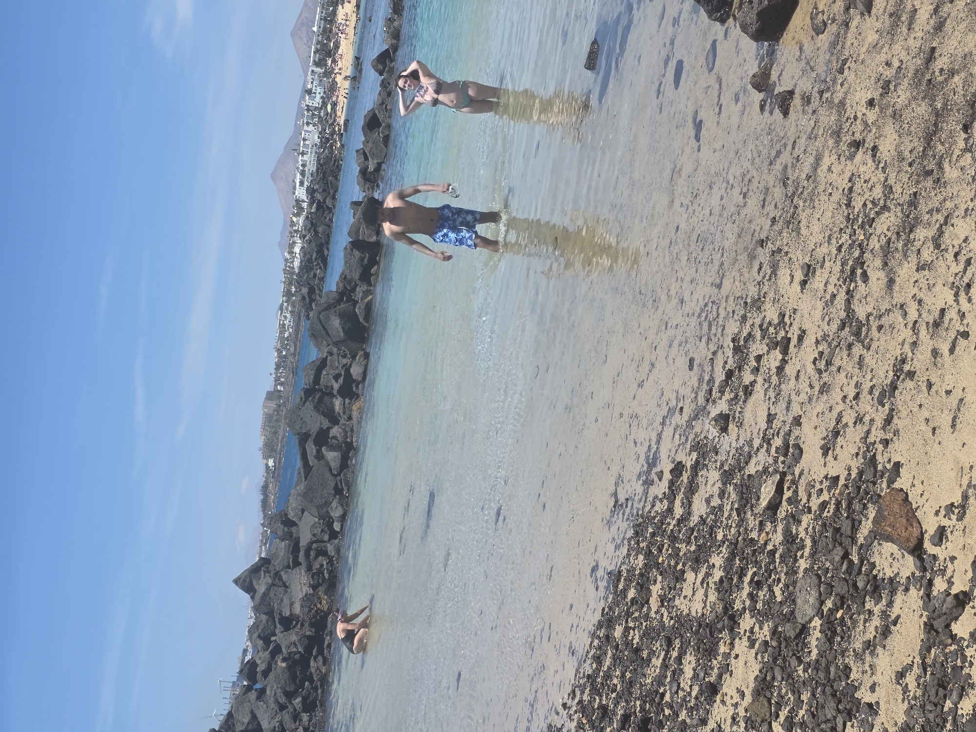 Three individuals are visible in shallow, clear ocean water. In the foreground left, a person in a black swimsuit and purple snorkel mask is partially submerged, leaning forward and reaching into the water. Further back and to the right, a shirtless person wearing blue patterned swim trunks stands in the water, holding a pair of goggles. Beside them, a person in a bikini stands with their hands on their head, facing the viewer. The background shows a rocky shoreline with dark volcanic rocks and a sandy beach dotted with buildings and mountains under a bright blue sky with scattered clouds. The scene appears to be in Costa Teguise, Spain, likely near a volcanic beach area.