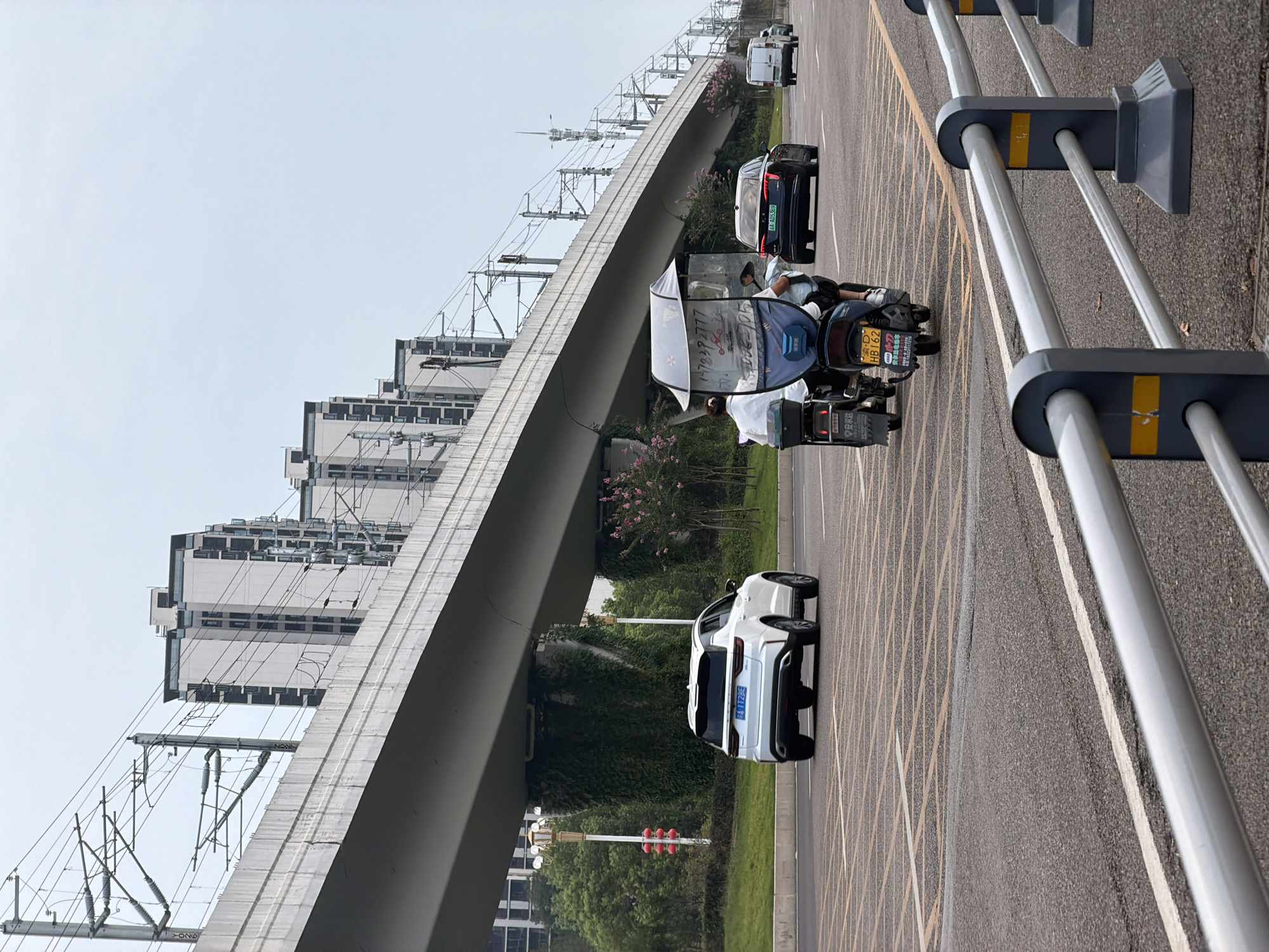 The image depicts a street scene in Jiangjin, China, under an elevated highway or railway overpass. A white SUV is driving on the road, followed by a scooter with two passengers. In the distance, several cars and a white van are also visible. The overpass is supported by columns covered in green ivy, and tall buildings can be seen in the background. The sky is overcast. A metal railing is in the foreground.