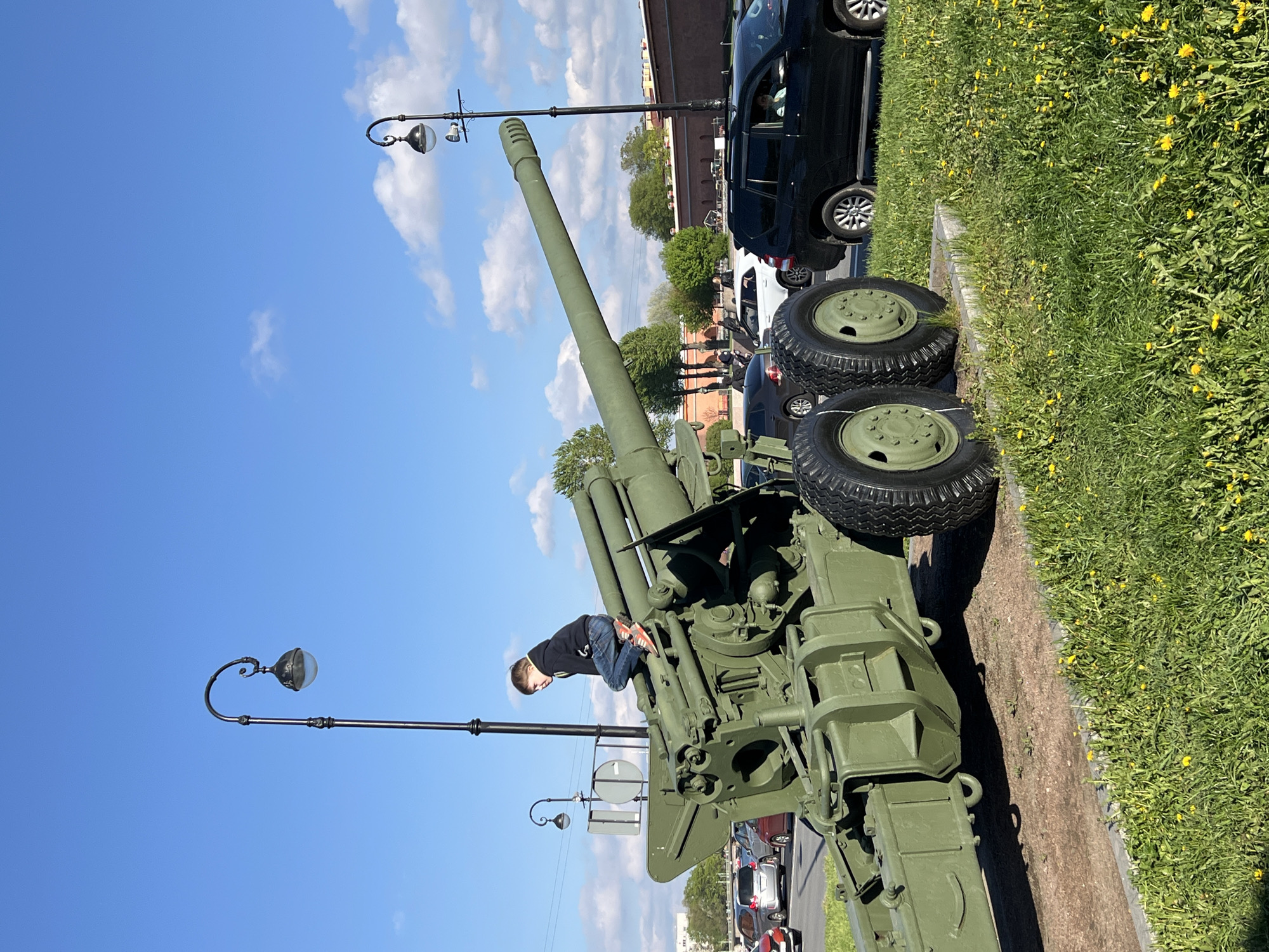 A boy is climbing on an olive green artillery gun. The gun is on display next to a sidewalk bordered by green grass with small yellow flowers. Parked cars are visible next to the grass and sidewalk, and there is a brick building in the background. The sky is blue with scattered white clouds, and there are black lamp posts next to the gun. Based on the architecture of the brick building and the style of the lamp posts, this location appears to be in Saint Petersburg, Russia.