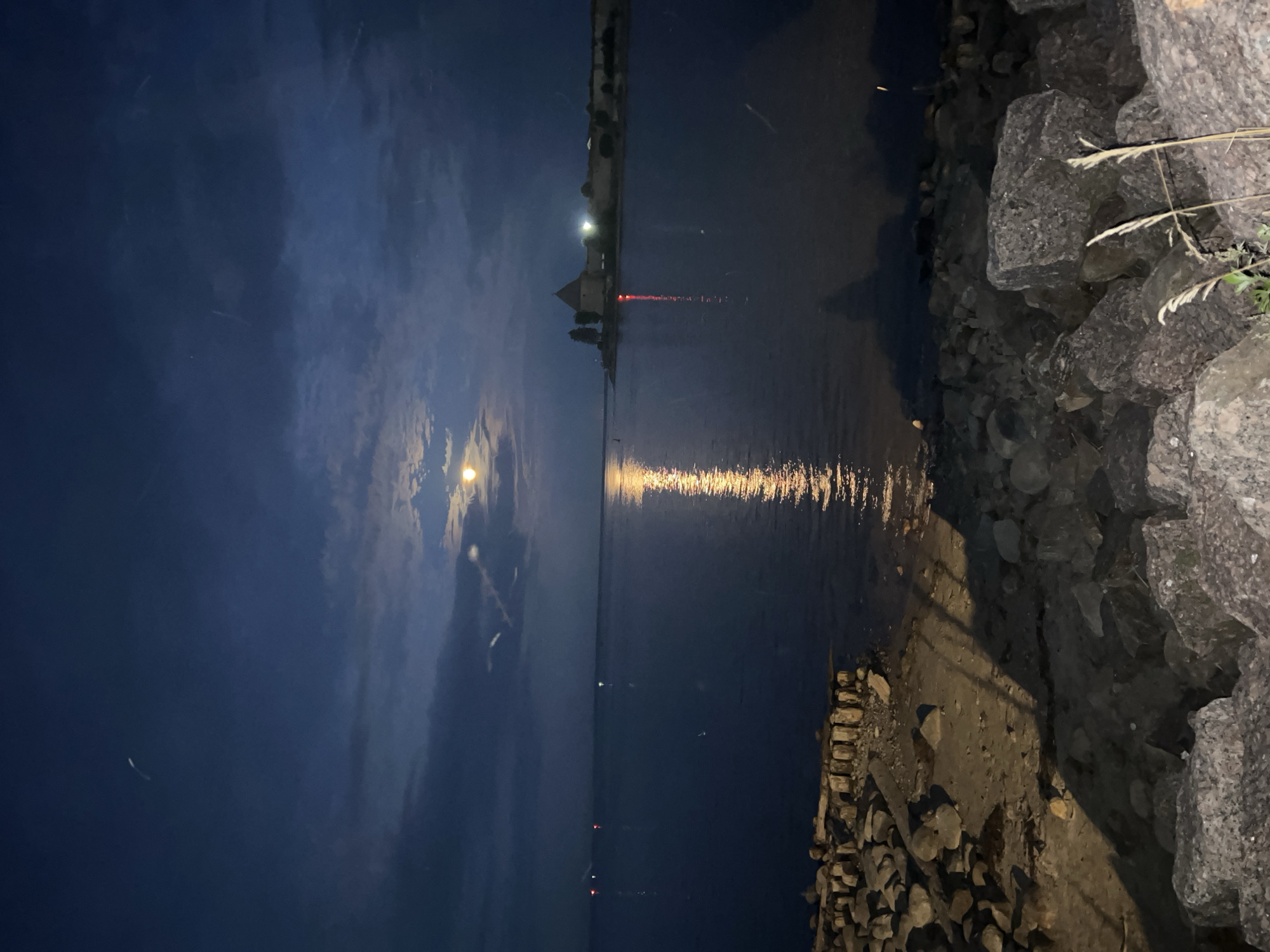 The image depicts a nighttime scene on the Neva River near the city of Imeni Morozova, Russia. The sky is dark blue with scattered clouds, and the moon is partially obscured by clouds, casting a bright reflection on the water. On the left bank of the river, there is a rocky shore with some wooden structures and logs. On the right bank, a fortified structure, possibly a fortress or castle, is visible in the distance, illuminated by lights. The water is calm, with several buoys or markers visible as small lights in the distance. The overall atmosphere is serene and somewhat mysterious. There are no people visible in the image.