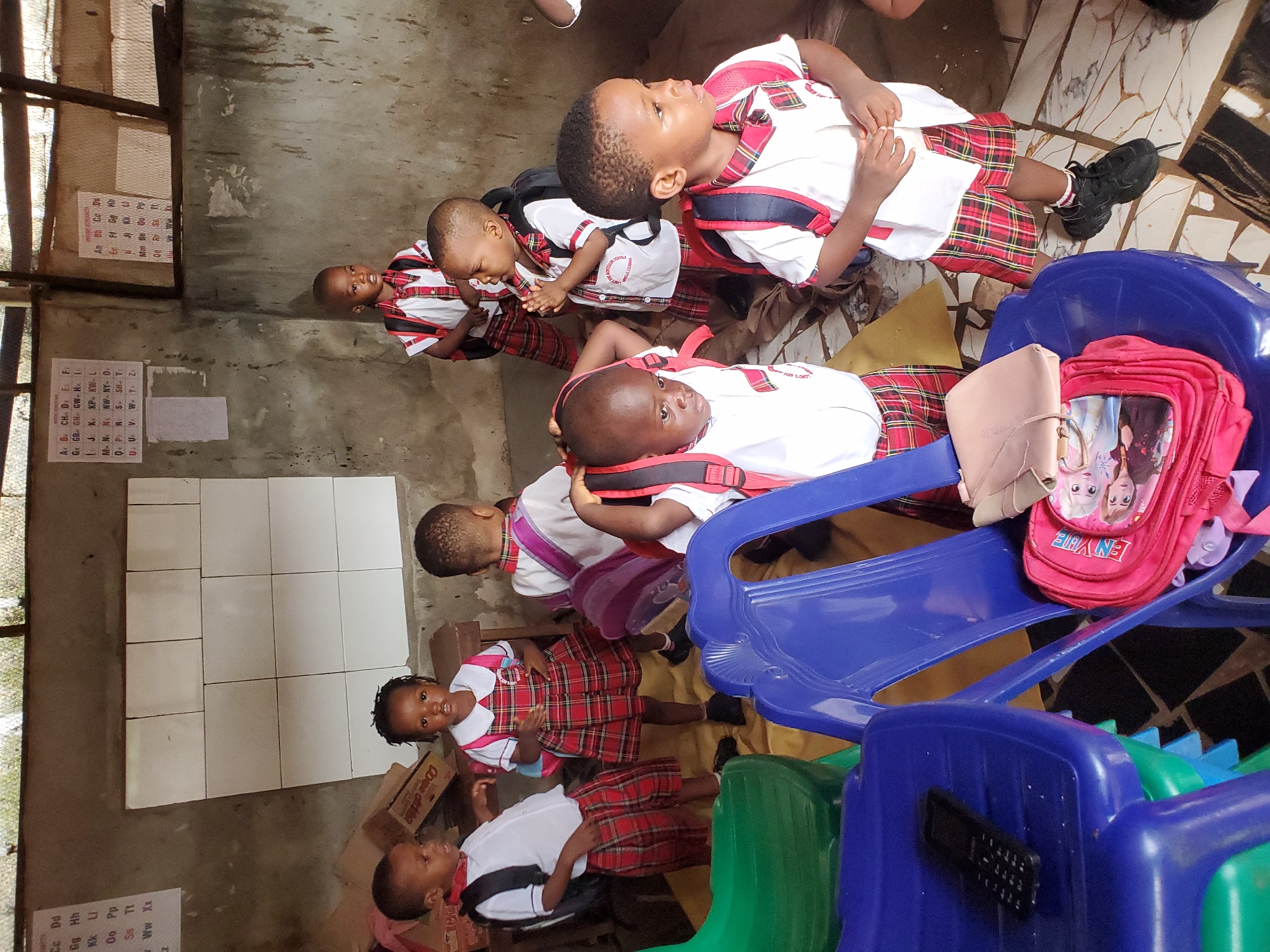 The image depicts a group of young children in school uniforms, likely students, positioned in what appears to be a classroom setting in Ukwu-Abwa, Nigeria.
The children are wearing matching school uniforms consisting of white shirts and red and black plaid bottoms. Some have backpacks on, varying in color and style. Some of the children are sitting on or near plastic chairs, primarily in blue and green. A remote control sits on one of the chairs.
The classroom setting includes educational materials such as alphabet charts on the wall. The walls themselves appear to be a combination of concrete and tiled surfaces. The overall setting seems simple and perhaps not very spacious.