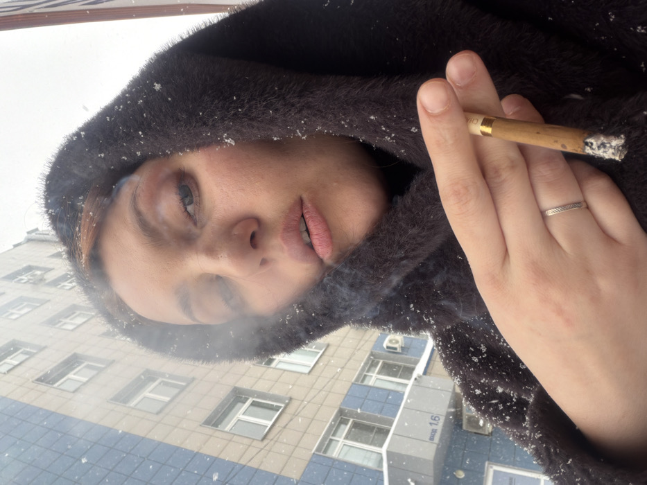 An individual, likely female, is depicted outdoors in Chaikovsky, Russia, during a snowfall. The subject, viewed from a low-angle close-up, has light skin, visible light brown hair, and a small stud piercing in the left nostril. She is wearing a dark, possibly faux fur, hooded garment dusted with snowflakes. Her left hand, adorned with a simple silver-colored ring on the ring finger, holds a lit cigarillo with a white filter between the thumb and index finger. Smoke emanates from the cigarillo and around her face, indicating an exhalation. In the background, a multi-story building features light-colored panels interspersed with vertical blue-paneled sections and multiple windows. An exterior air conditioning unit is visible on the building. A sign on the building's ground floor clearly displays the Cyrillic text "ВХОД 1.6", translating to "Entrance 1.6".