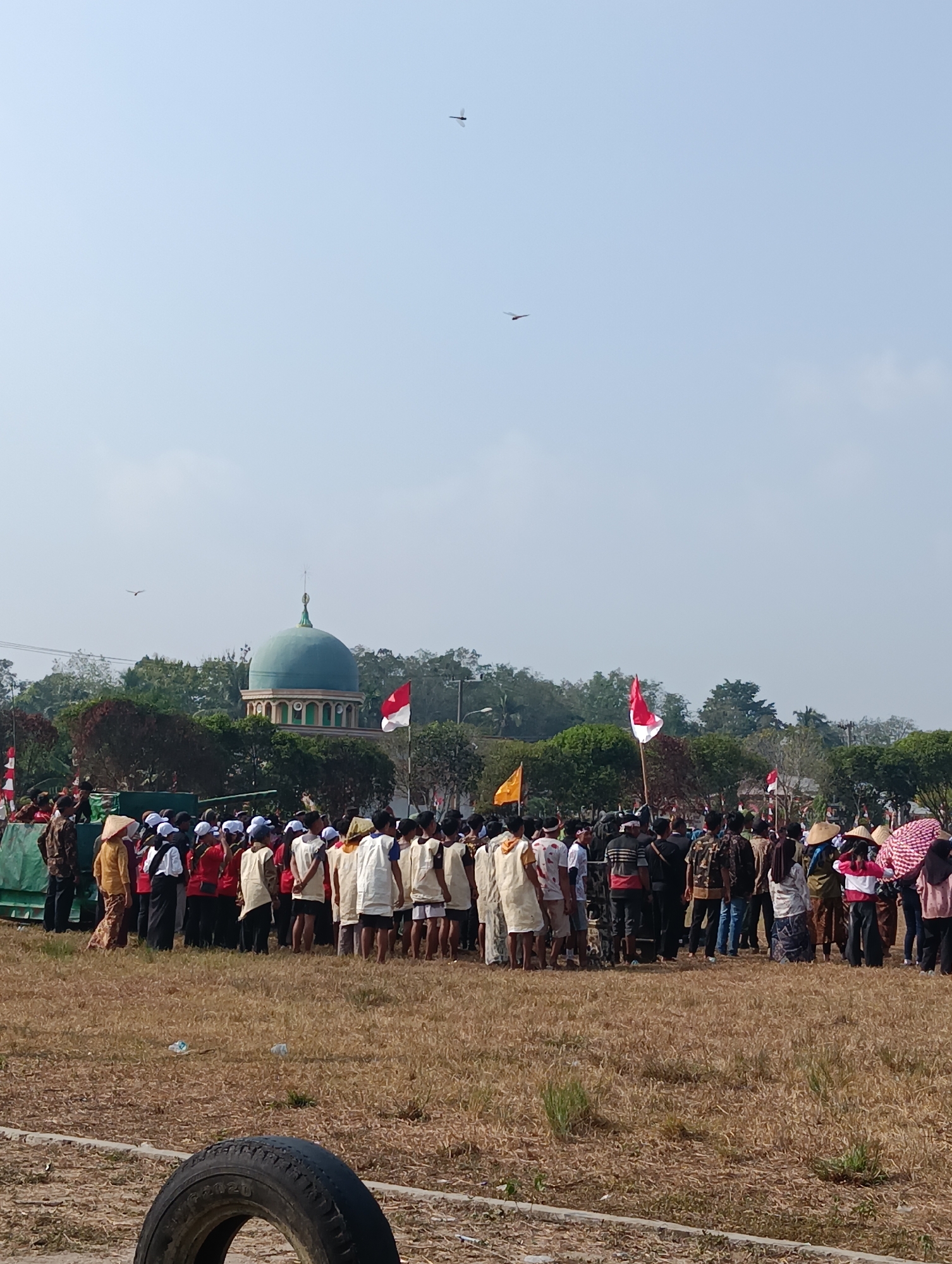 This is a full shot taken from a slightly low angle, outdoors, on a bright, clear day. The image depicts a large gathering of people, likely a ceremony or event, in an open field. In the background, a distinctive teal-domed building, possibly a mosque, is visible, surrounded by lush green trees. The Indonesian flag, a bicolor of red and white, is prominently displayed, along with other flags. The crowd consists of men, women, and children, many wearing traditional Indonesian clothing, such as conical hats and batik. Some are wearing white or light-colored uniforms, suggesting participation in a group activity or school event. A few individuals are holding umbrellas, possibly for shade. The ground is covered in dry, brown grass, and there is a discarded tire in the foreground. Two small, dark shapes are visible in the sky, which appear to be insects or birds, one in the upper right quadrant and another in the mid-left. The overall atmosphere suggests a community event taking place in a rural or semi-rural setting in Pematang Jaya, Indonesia.