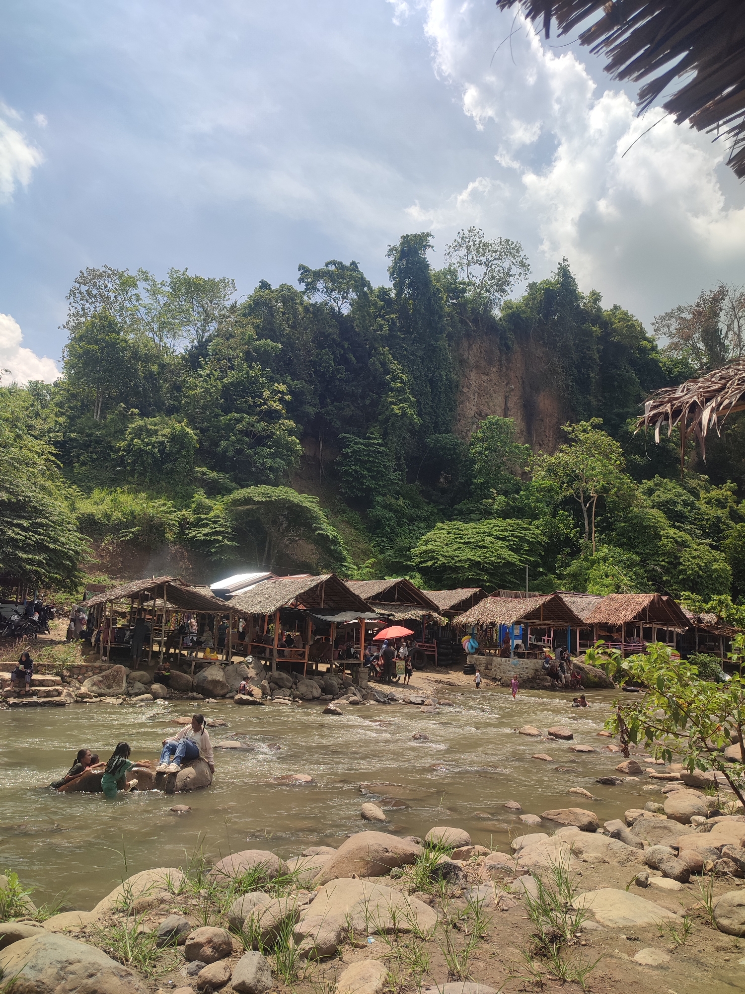In Pantonrayeuk Satu, Indonesia, a scene unfolds by a river with a rocky shore. A row of open-air bamboo and thatched-roof huts lines the riverbank, some with people seated inside or standing nearby. In the water, several individuals are present. On a large rock in the river, a young woman in jeans and a light-colored top sits, while two other young women, one in a green swimsuit and another partially submerged, interact with her. Further downstream, a few more people are visible in the water. The river flows over rocks and around submerged stones. The background features a steep, vegetated cliff face covered in lush green trees and foliage. The sky above is a mix of blue with scattered white clouds. The overall atmosphere suggests a recreational area by a river in a tropical, natural setting.
