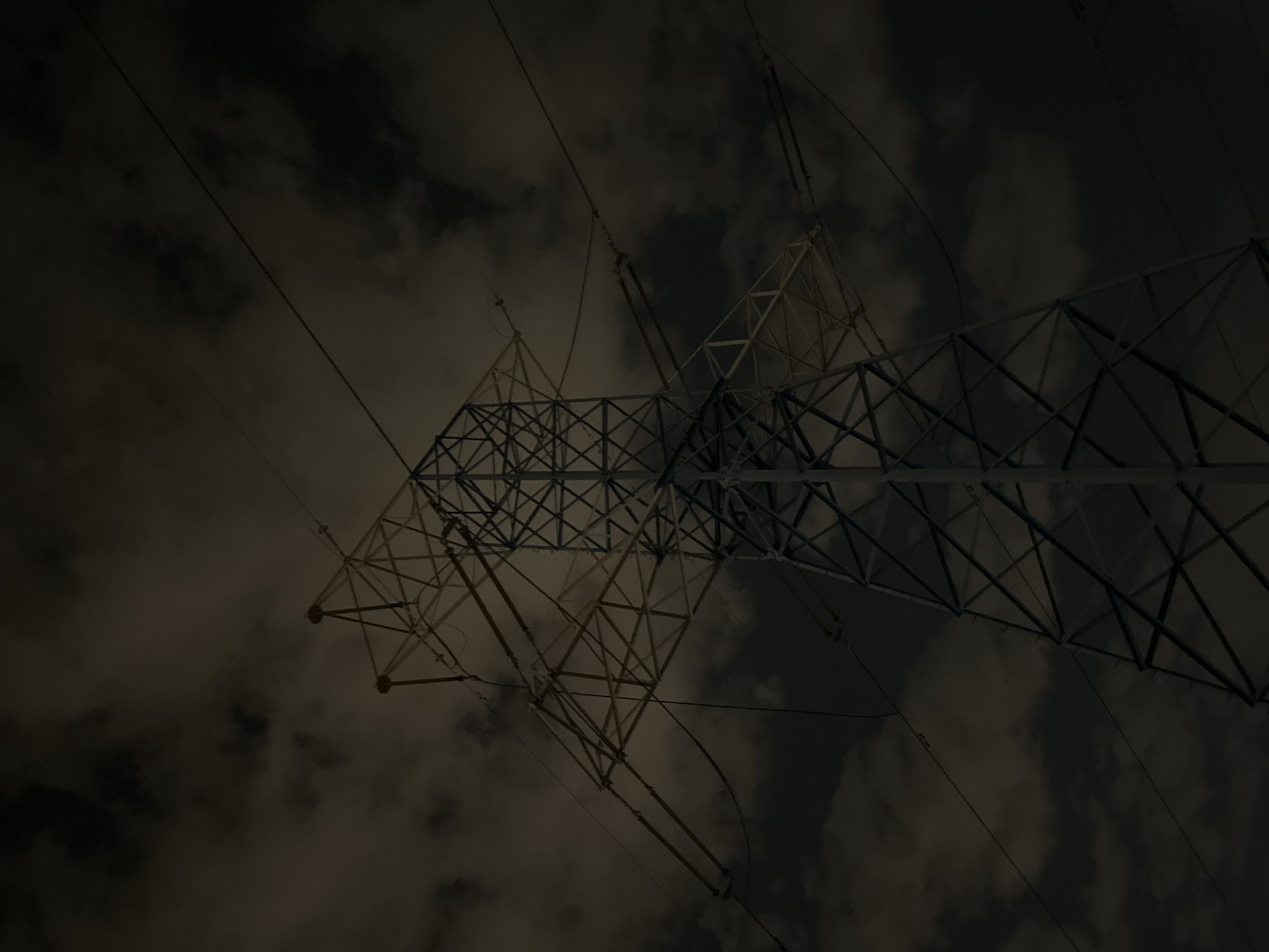 A low-angle shot captures an electricity pylon against a cloudy sky. The lattice structure of the pylon is visible, with a crisscrossing pattern of metal beams. Electrical wires extend from the pylon. The clouds are a mix of dark and light tones. There's no indication that this image is in Moscow, Russia.