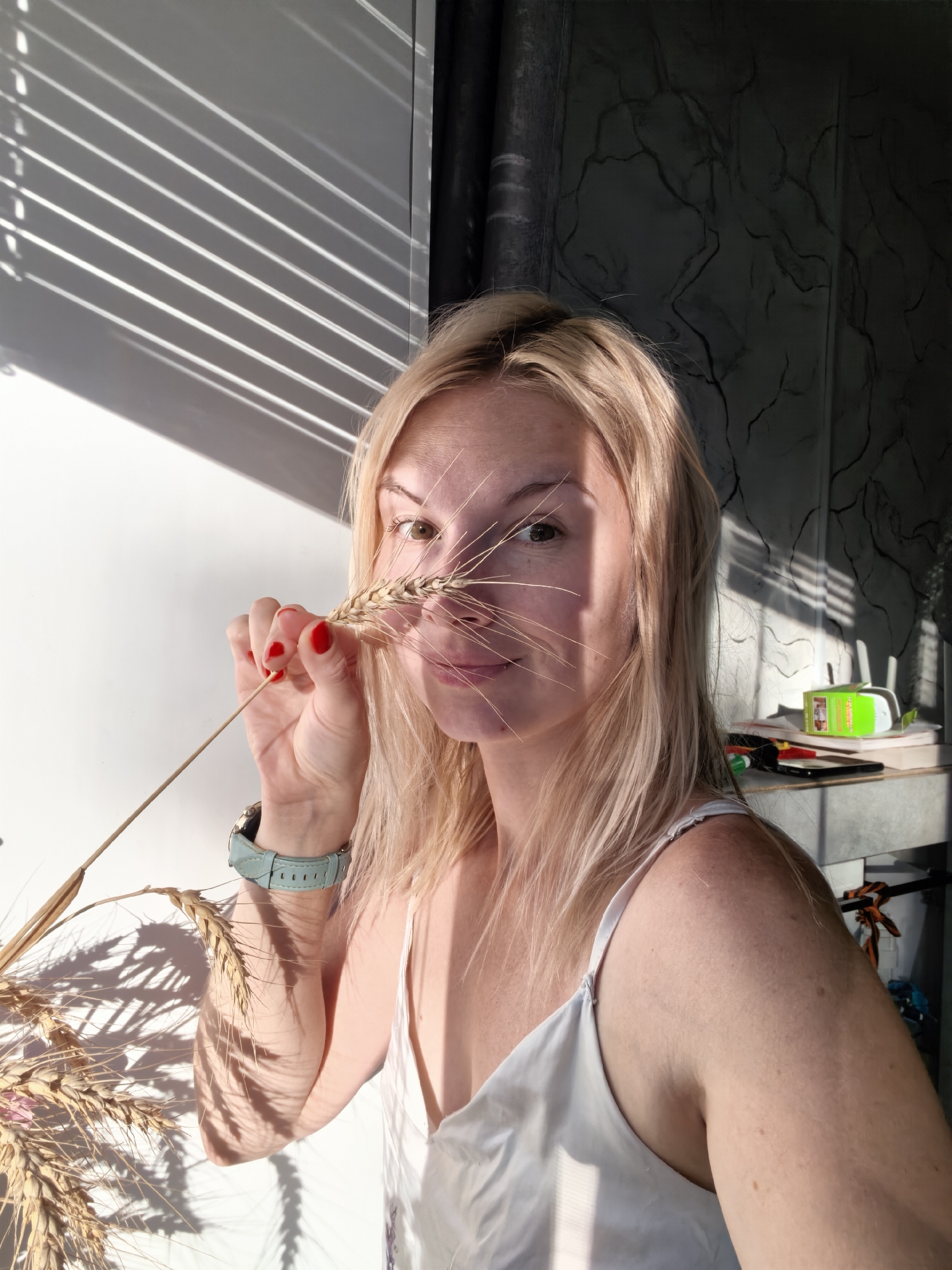 The image shows a blonde woman holding a stalk of wheat in front of her face. She is wearing a white camisole and a light blue watch on her left wrist. The background features a white wall with shadows from blinds, and a dark, textured wall on the right. On a shelf to the right, there are some objects including a green container, pens, and a mobile phone. The lighting suggests an indoor setting with sunlight streaming through blinds. The image was taken in the city of Sterlitamak, Russia.