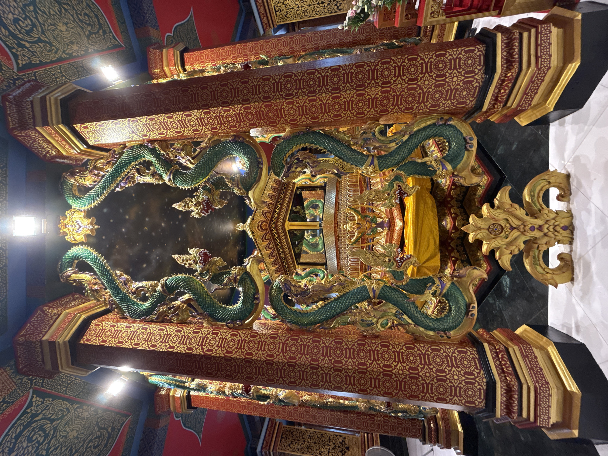 The image shows the ornate interior of a temple in Ban Khao Tang, Thailand. The central focus is a golden altar or shrine, draped with yellow fabric and adorned with intricate carvings. Flanking the altar are two massive, green, serpent-like sculptures with gold accents, each coiled and rearing upwards. These serpents are attached to large, red and gold patterned pillars. The ceiling is decorated with vibrant patterns and a starry sky motif, illuminated by bright spotlights. The overall scene is characterized by rich colors, elaborate decorative elements, and a sense of sacredness. There are no people visible in the image.