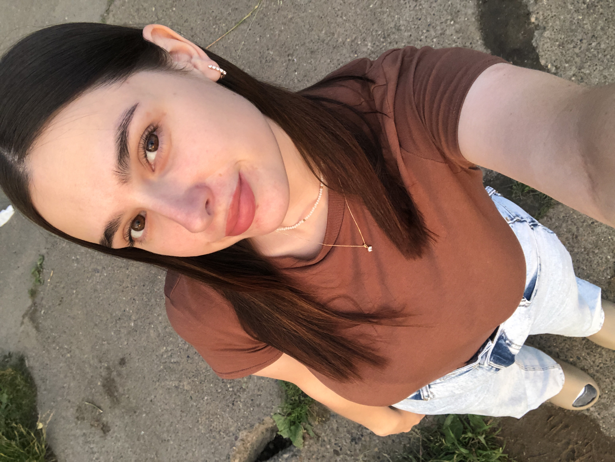 A young woman with dark hair is taking a selfie outdoors. She is wearing a brown t-shirt, light-wash jeans, and tan slides. She has on a delicate pearl necklace with a small pendant and a sparkling earring in her left ear. The woman has large, expressive brown eyes and her lips are painted a soft pink. She is smiling slightly as she looks at the camera.
The background appears to be an asphalt surface, possibly a sidewalk or road, with some patches of grass visible. The lighting suggests it is either early morning or late afternoon, with the sun casting a warm glow. The overall mood is casual and relaxed. There is no visible text in the image. The location is Chaikovsky, Russia.