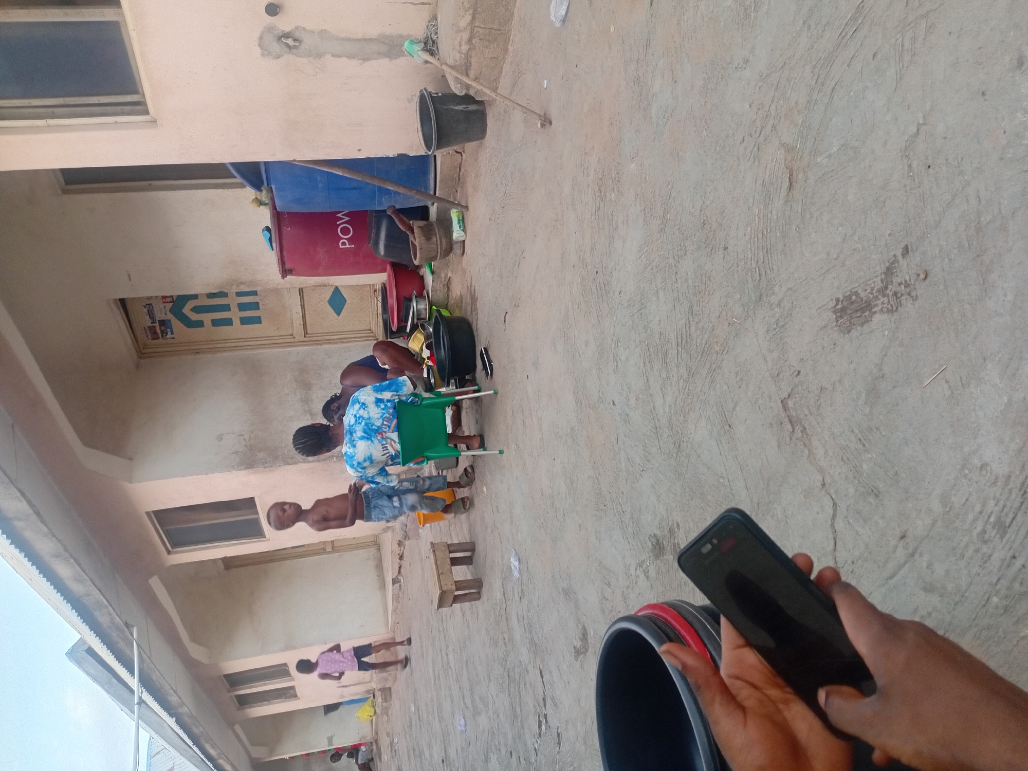 This image captures an outdoor scene, likely a courtyard or a passage between residential buildings, illuminated by natural daylight. The overall setting is characterized by concrete ground and light-colored, possibly peach or beige, painted walls.
In the immediate foreground, a pair of dark-skinned hands holds a black smartphone, with a black plastic bucket featuring a red rim partially visible on the right.
The midground is populated with several individuals and household items. An adult, seen from behind, is seated on a green plastic chair, wearing a blue and white tie-dye patterned top. Nearby, a young boy stands, shirtless and wearing denim shorts and sandals. Further into the scene, a smaller child in a pink top and dark shorts is visible. Other figures are present in the background, partially obscured, some appearing to be engaged in an activity involving various containers.
Numerous domestic objects are scattered across the concrete ground. These include several black plastic pots or bowls, a wooden mortar and pestle, and a green plastic bottle. A red cooler, with the word "POW" clearly visible vertically on its side, stands next to a large blue plastic barrel or container. A broomstick leans against one of the walls, and a small wooden stool is placed on the ground.
The background features the facades of two buildings. They have windows and doors; one central door displays a distinctive blue and white geometric pattern. The sky is visible in the upper portion of the frame, confirming the outdoor setting. The visible text "POW" is located on the red cooler.