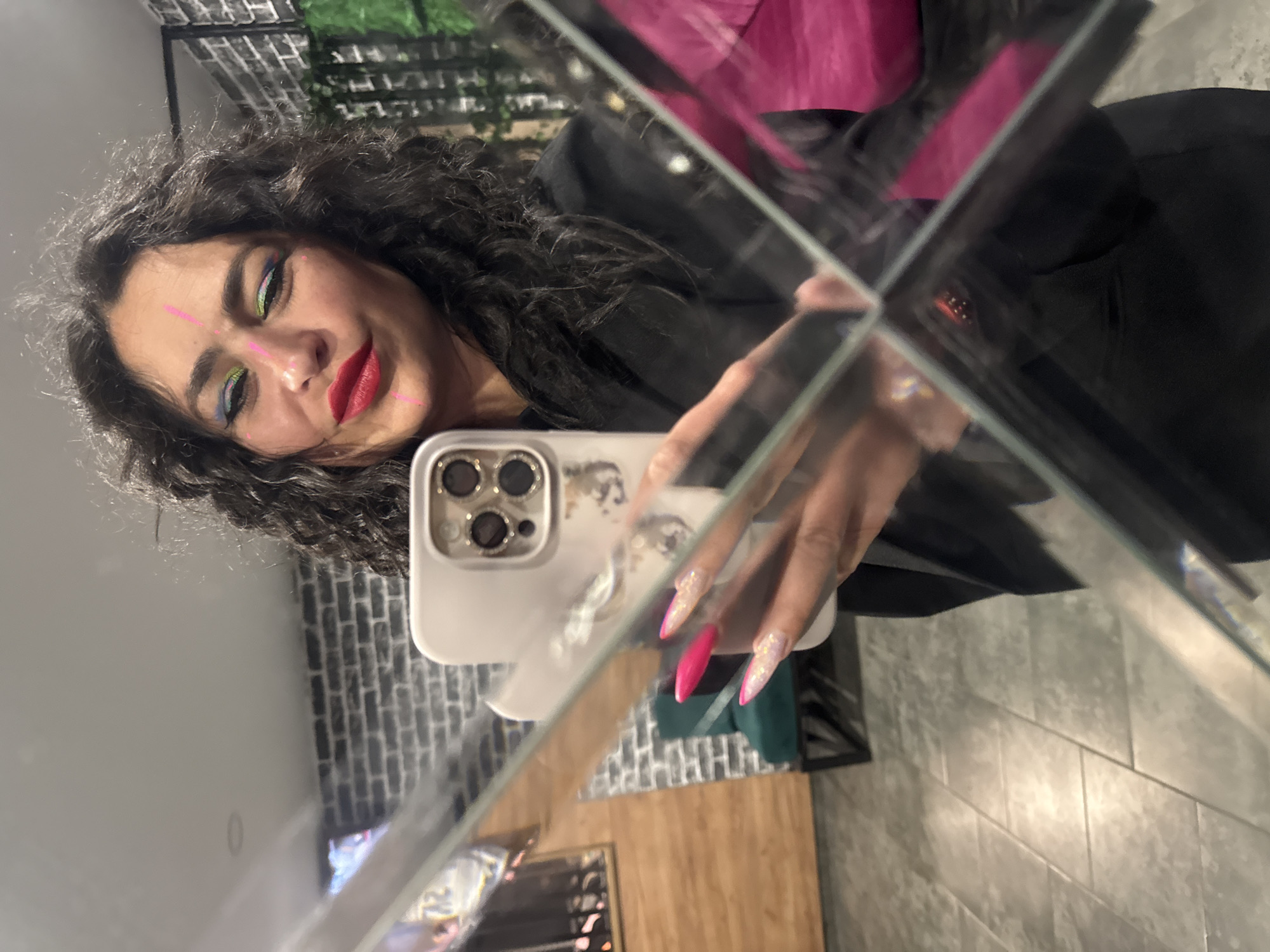 A dark-haired woman with colorful makeup and red lipstick is taking a selfie in a mirror. She holds a white smartphone, decorated with a pattern of eyes, in her right hand. She is wearing a black blazer. Her nails are manicured with glitter and pink tips. The reflection shows a room with grey brick walls. Outside the mirror is an area with green foliage and a glimpse of the room's interior: a wooden surface and grey tile. No specific location details for Chaikovsky, Russia, are discernible from the image.