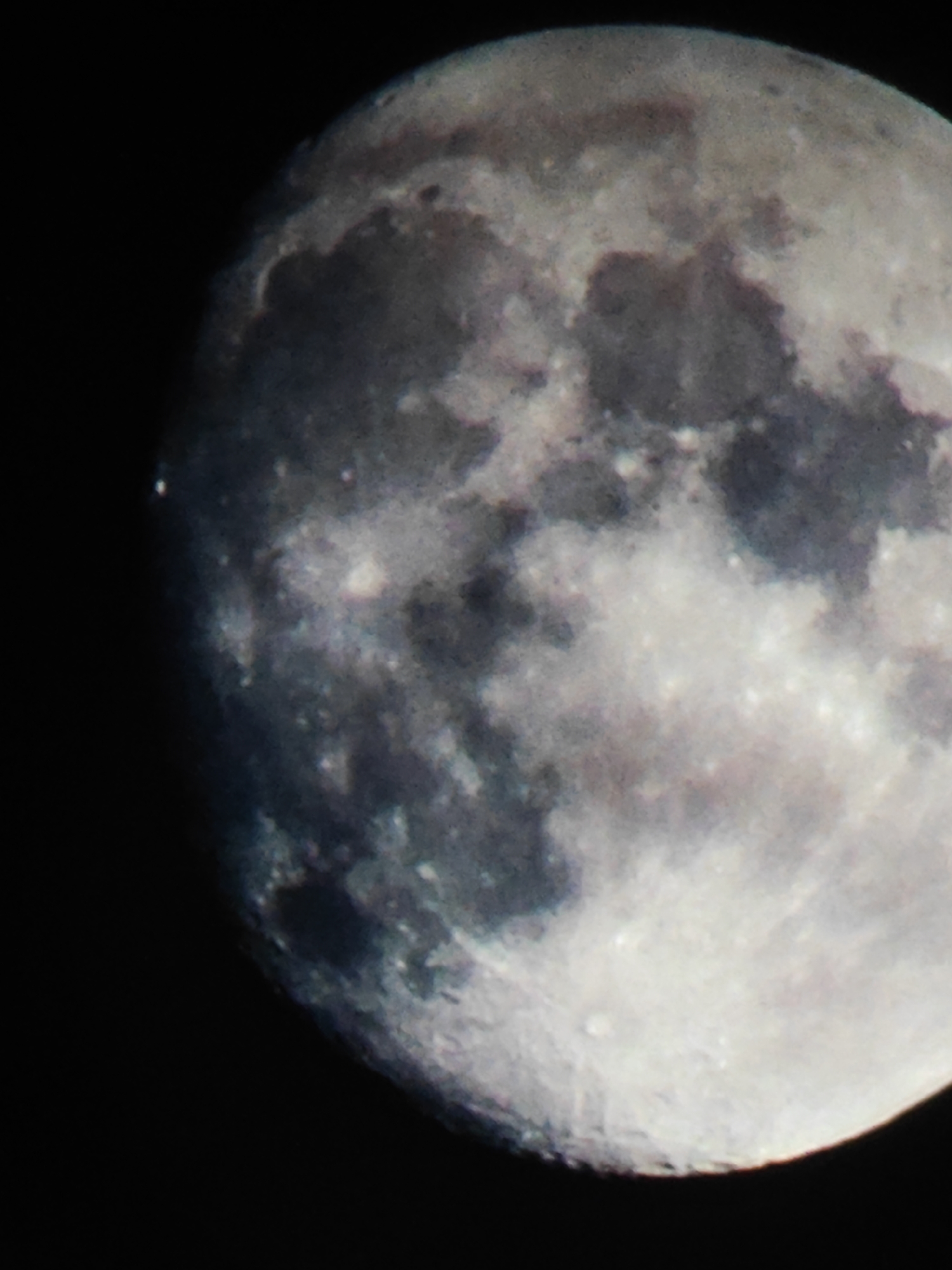 The image shows a close-up view of the moon against a black background. The moon is in a waxing gibbous phase, with a significant portion illuminated. The surface is textured with craters and maria (dark, smooth plains). There is a small bright spot visible on the left edge of the moon. The image doesn't offer sufficient context to establish a location in Chaikovsky, Russia, or any information about people, actions, interactions, or the environment.