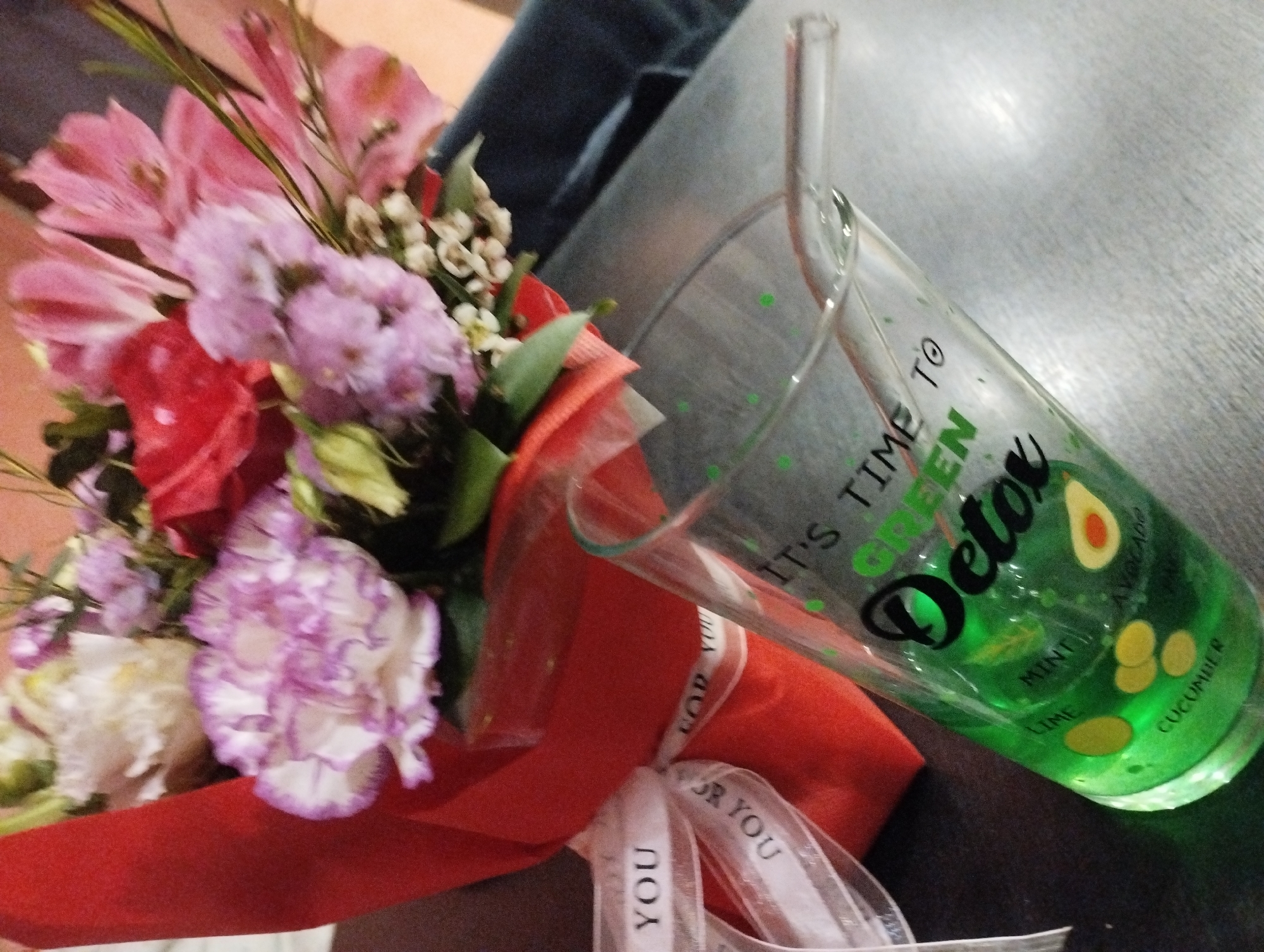 The image shows a close-up of a bouquet of flowers and a glass of green juice on a dark surface. The flowers are a mix of pink, red, and purple hues, wrapped with a red and white ribbon. The juice glass is transparent with a straw and contains a green liquid with images of avocado, mint, lime, and cucumber. The text on the glass says, "It's time to Detox." The background suggests an indoor setting. There is no indication of the location in Burgas, Bulgaria. No people are visible.