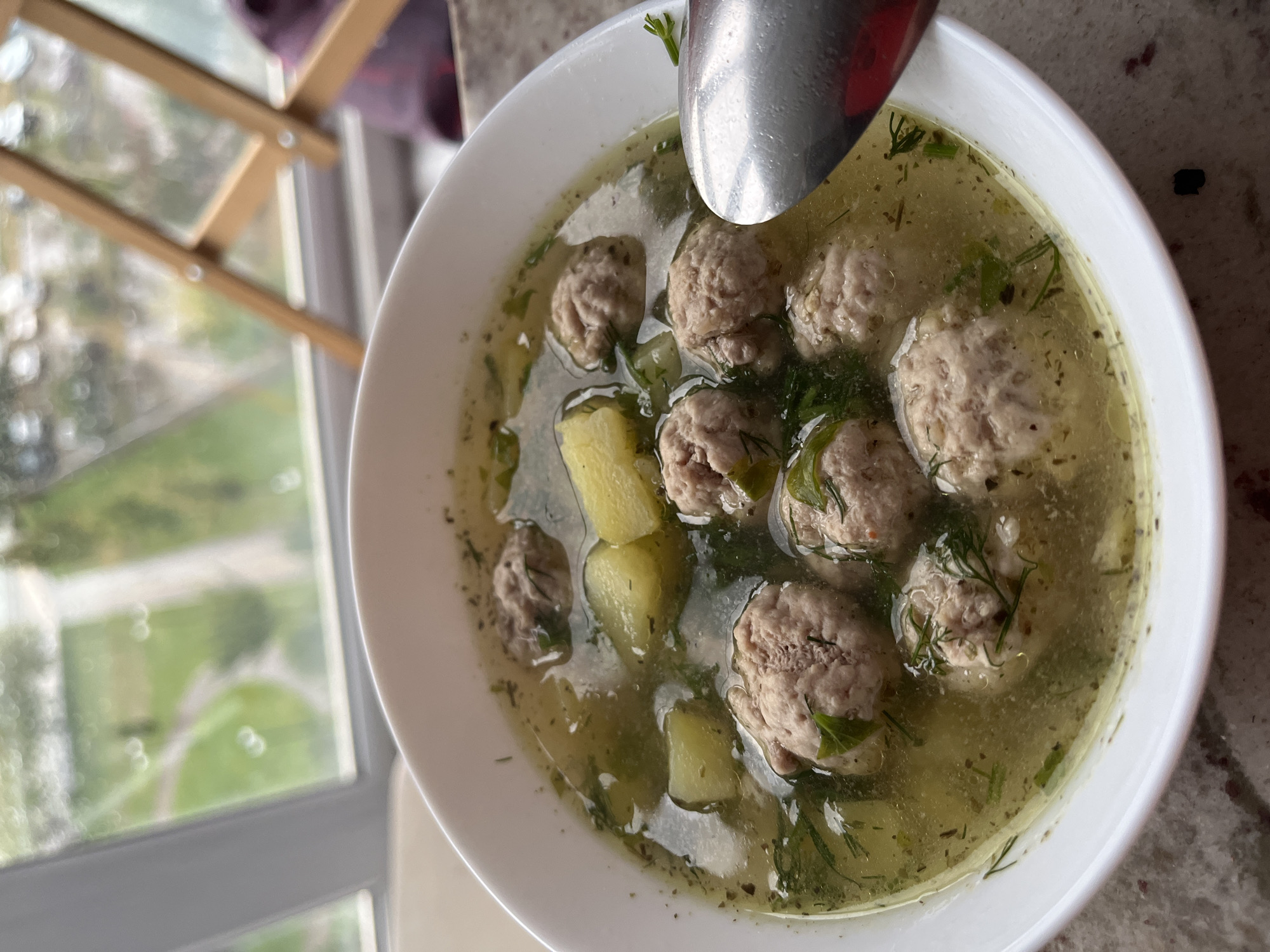 A white bowl of soup filled with meatballs, potato chunks, and dill sits on a granite countertop. A silver spoon is partially visible on the right side of the bowl, ready to scoop the soup. In the background, a window offers a blurry view of a green landscape with trees and a winding path, suggesting an urban setting. A wooden folding chair is also visible in the background, partially obscuring the view. The overall lighting is natural, possibly from the window.