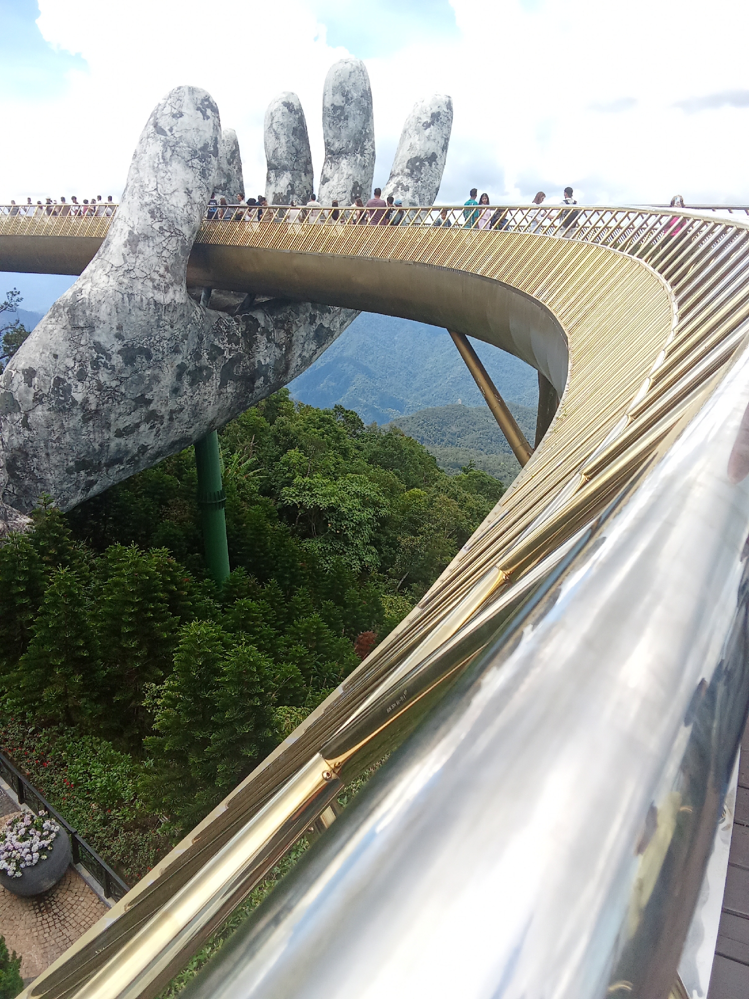 A surreal, massive concrete hand emerges from a lush, green mountainside, with a golden bridge curving gracefully upwards from its palm. The bridge is supported by a large green pillar attached to the underside of the hand. The hand's textured surface, with its gray and white marbling, contrasts with the smooth, metallic sheen of the bridge's railing, which occupies the foreground of the image. Tourists are seen walking along the bridge, their figures small against the monumental scale of the hand. The background reveals a misty, mountainous landscape, suggesting a high elevation. The scene is set in the Ba Na Hills of Đà Nẵng, Vietnam, a popular tourist destination known for its unique architecture and breathtaking views.