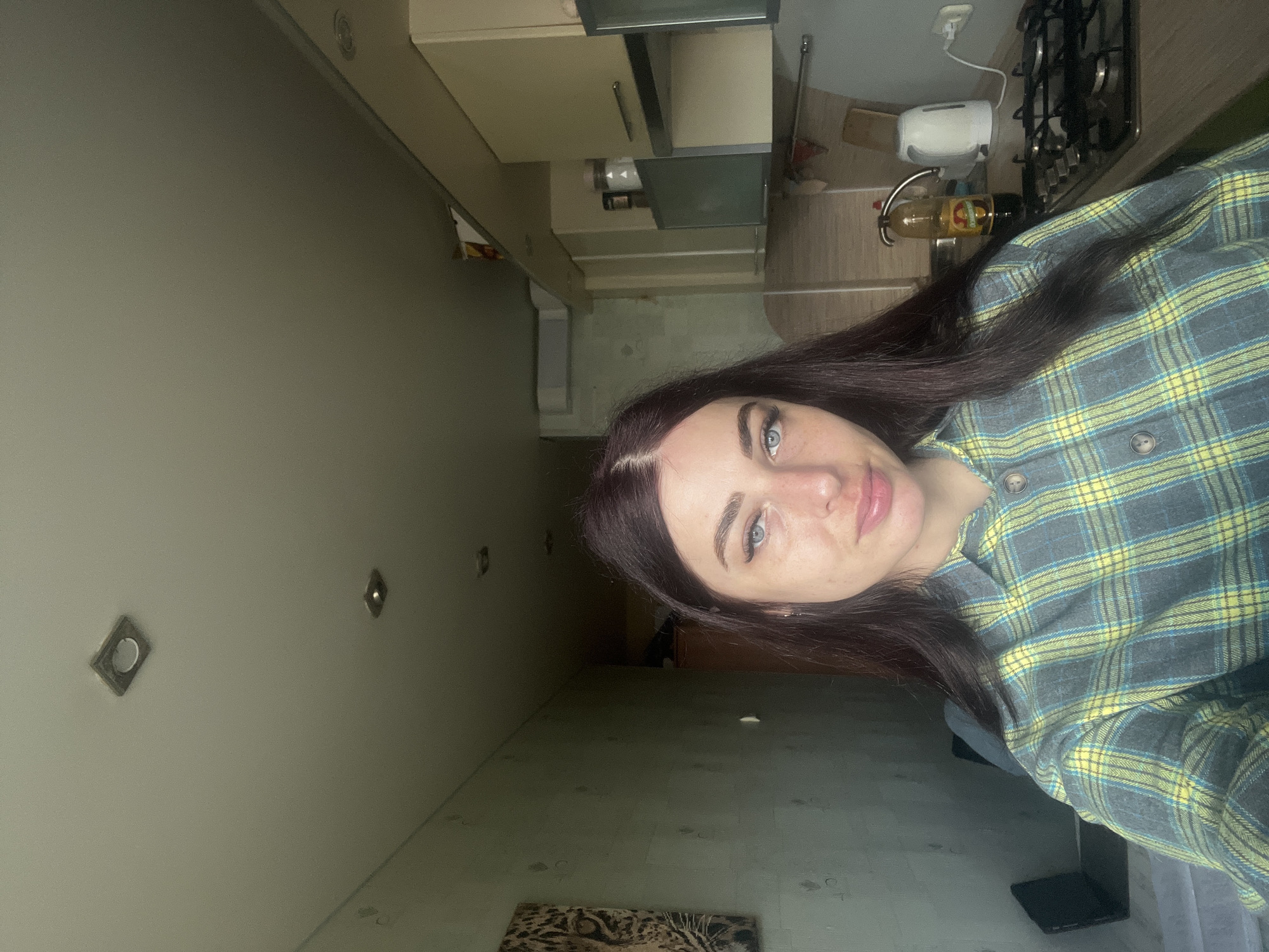 A selfie captures a young woman indoors, likely in a kitchen or dining area within a residence in Chaikovsky, Russia. The woman, with long, dark hair and blue-green eyes, looks directly at the camera with a neutral to slightly contemplative expression. She is wearing a grey and yellow plaid button-up shirt. Her face occupies the right side of the vertically oriented frame.
The background on the left features a plain, light grey wall with two square recessed ceiling lights visible. To the right and behind her, kitchen elements are prominent. These include light-colored cabinets, some with dark glass panels, and a countertop. On the countertop, a black gas stovetop with multiple burners is visible, along with a white toaster and a clear glass bottle, possibly containing oil or a beverage. The bottle has a dark label, but no discernible text.
The floor, visible towards the bottom of the image, appears to be a light-colored surface, possibly linoleum or wood, with various dark scuff marks or stains. A portion of a textured rug or mat, with what looks like an animal print (possibly leopard), is partially visible in the bottom-left corner. The lighting in the room appears to be from artificial sources and is somewhat dim in areas, suggesting it could be daytime indoors or evening. No external windows are visible. The overall scene conveys a casual, domestic setting.