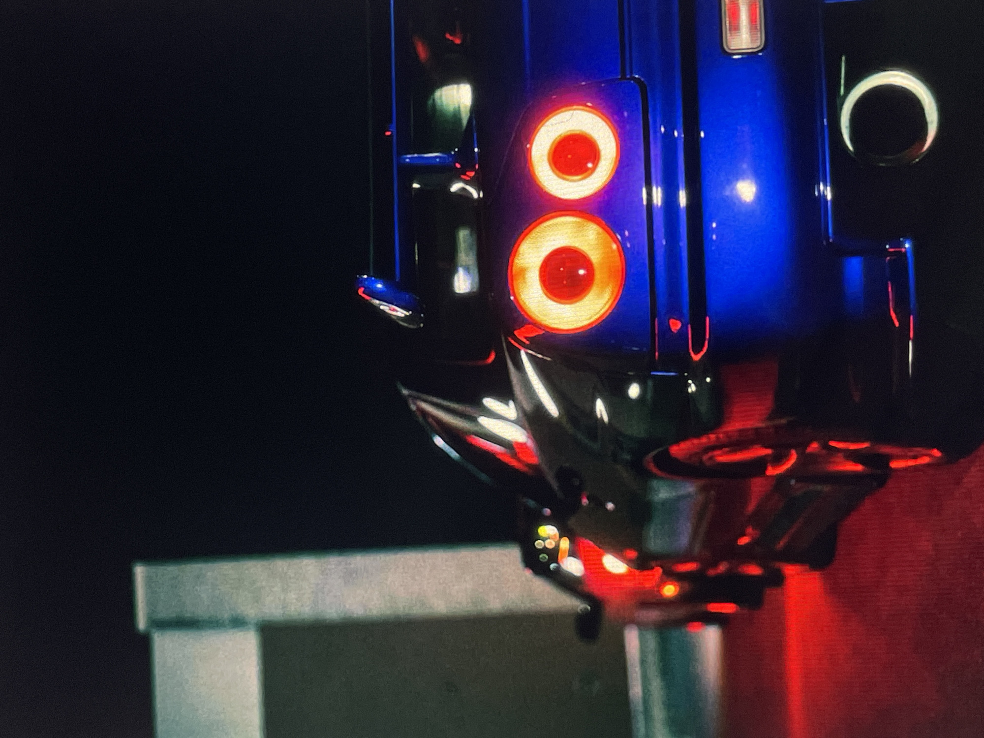 The image shows a close-up view of the rear of a dark blue sports car, likely a McLaren, with its taillights illuminated. The car is elevated on a lift or stand, as visible by the support structure below. The background is mostly dark, except for a gray horizontal surface and red illumination. There is no indication of location.