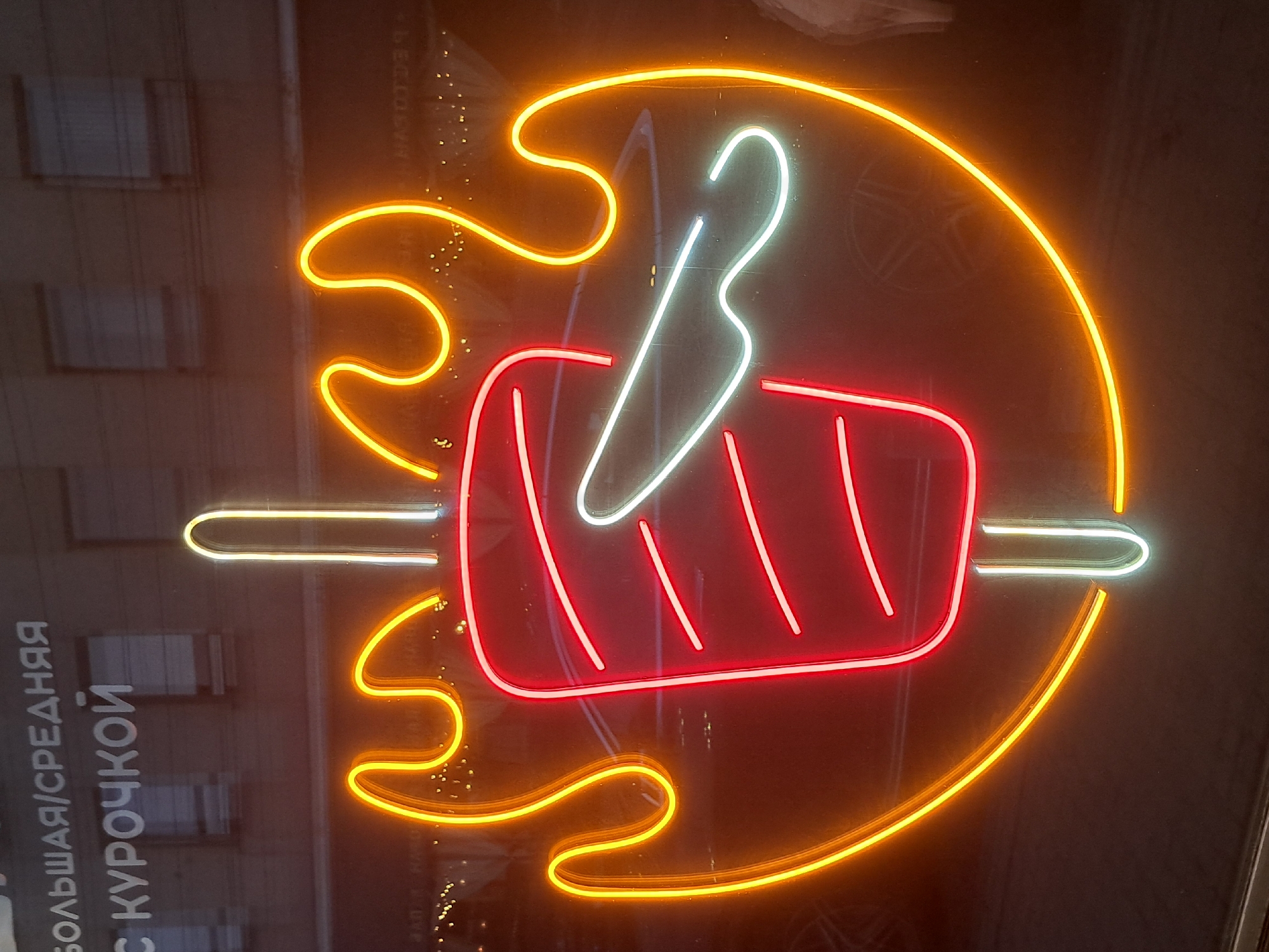 The image shows a neon sign outside a shop in Irkutsk, Russia. The sign depicts a rotisserie kebab with a knife slicing it, surrounded by flames. The neon colors are red, yellow, and white. Text in Russian is visible above the sign. The background appears to be a dark building with windows. The scene is a street view, likely at night or in low light conditions, with the neon sign being the primary light source. The overall impression is that of a food establishment, possibly a kebab shop or restaurant.
