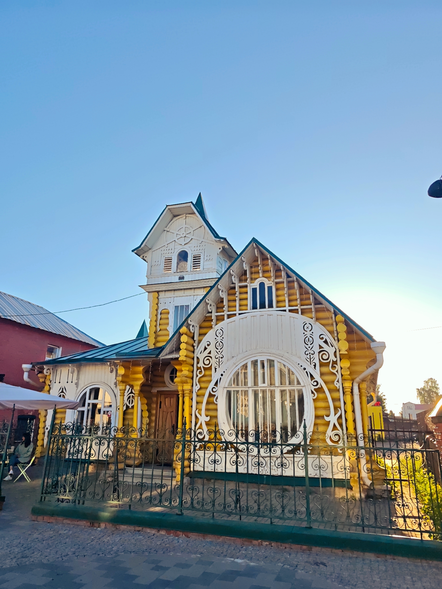 In Kimry, Russia, a brightly painted wooden house with intricate white trim stands under a clear blue sky. The house features a distinctive round window framed by ornate white carvings and a tall, multi-tiered tower. A green metal fence with decorative scrollwork encloses the property. To the left, a red building with a metal roof is visible, and a white umbrella shades an outdoor seating area where a person sits alone. The sun casts long shadows, suggesting it is either early morning or late afternoon. The scene appears to be on a street with a cobblestone or patterned paving.
