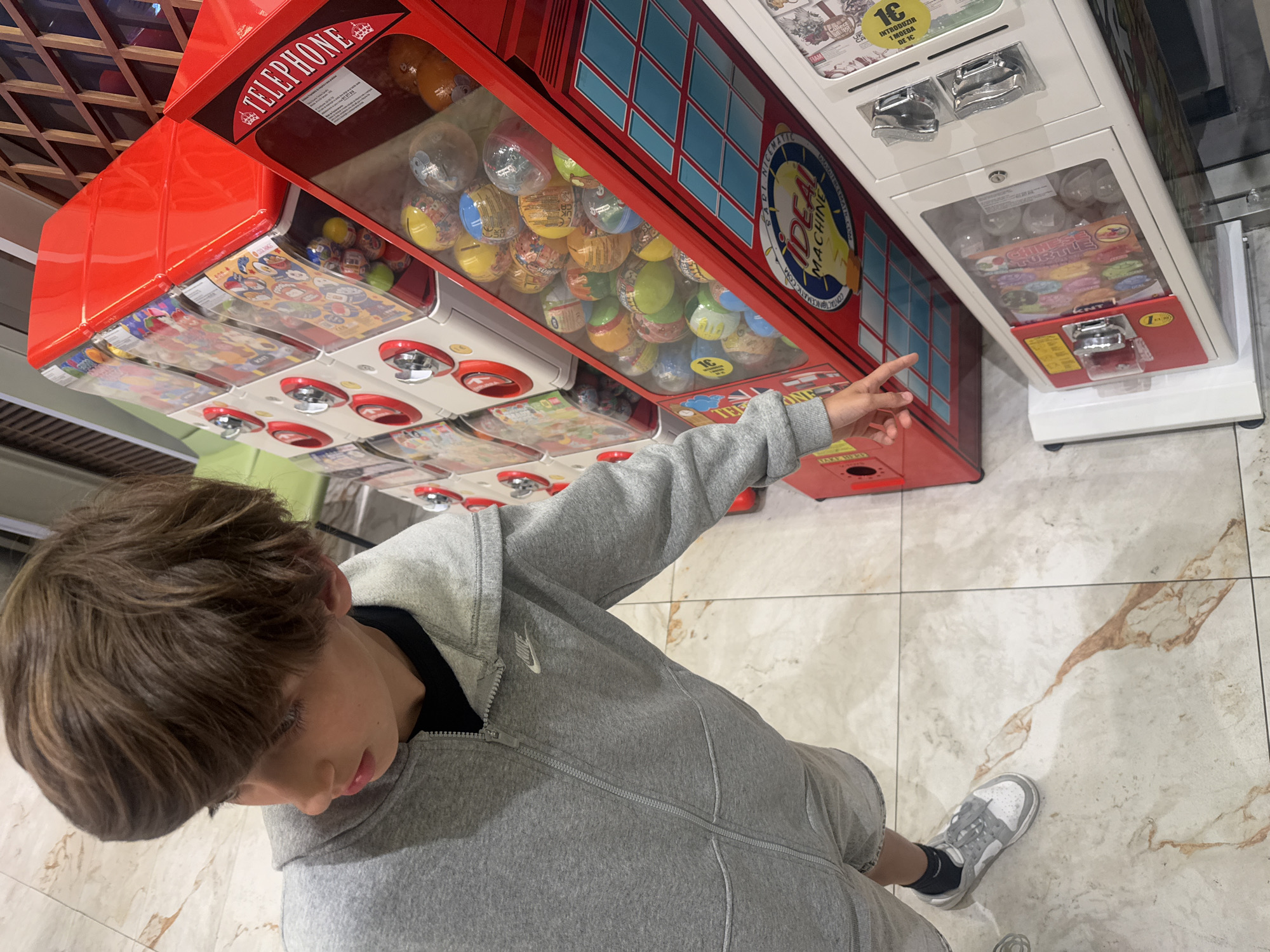 The image shows a young person, viewed from above, pointing towards a coin-operated toy vending machine. The machine is shaped like a British telephone box and is filled with small toy-filled capsules. Next to it, there is another vending machine. The person is wearing a gray Nike zip-up hoodie, gray shorts, black socks, and gray and white sneakers. The floor is tiled. A partial view of a ceiling is seen in the upper left. Based on the machines and layout, this could be located in a shopping center or arcade in Vila Nova de Gaia, Portugal.