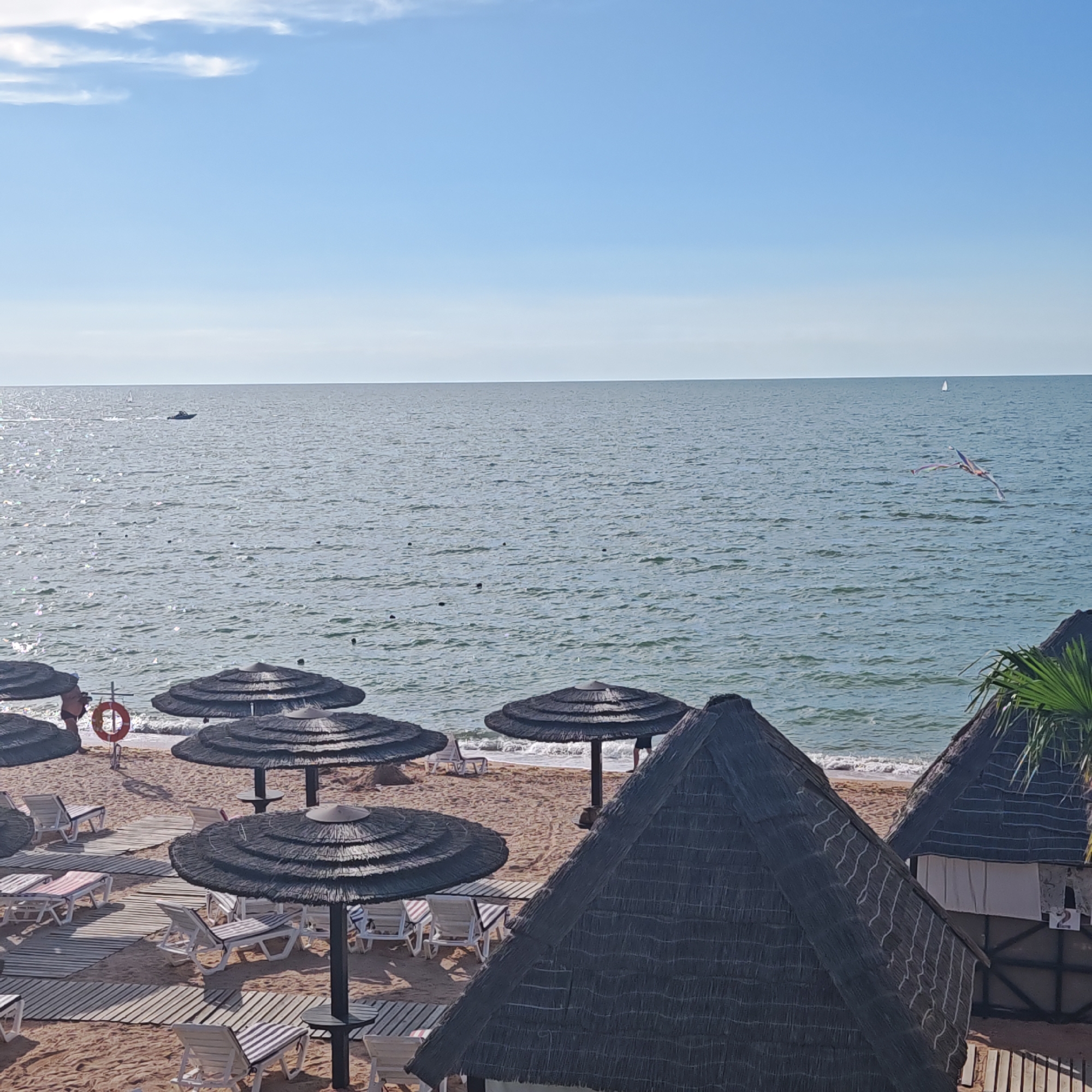 This media file depicts a sunny day at a beach resort. The scene is set against a clear blue sky with a few wispy clouds and the vast expanse of the sea. The water is calm with gentle waves lapping the shore.
In the foreground, there are several thatched umbrellas providing shade over empty lounge chairs arranged on wooden walkways laid out on the sandy beach. Some lounge chairs are occupied by people. On the beach, a red life preserver hangs on a pole, and a few people are visible wading in the shallow water.
Further out in the sea, a motorboat is creating a wake, and a small sailboat can be seen in the distance. A bird, possibly a kite, is soaring gracefully in the sky over the water. In the background, there are some small huts with thatched roofs, and one of them has the number "2" visible.
The overall atmosphere is peaceful and relaxing, suggesting a typical summer vacation day. The sunlight creates a shimmering effect on the water and casts shadows on the sand. The location is identified as Мысовское сельское поселение, Ukraine.