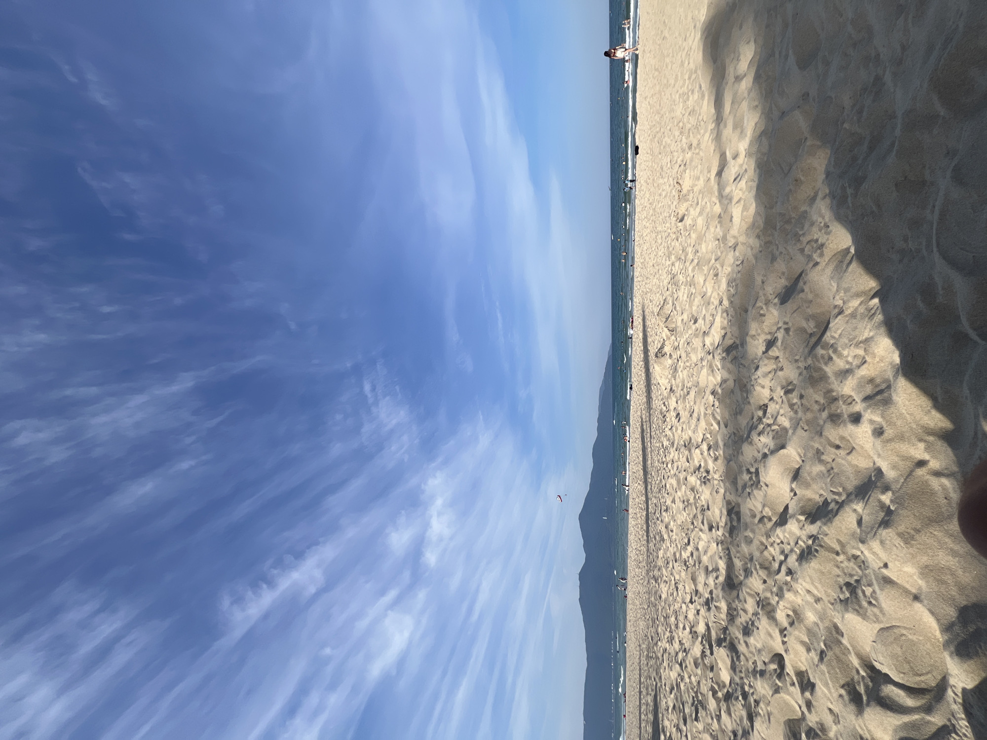 The image depicts a sunny day at a beach in Da Nang, Vietnam. The foreground is dominated by soft, rippled sand, casting shadows that suggest the sun is positioned to the left and slightly behind the viewer. In the mid-ground, the ocean meets the sandy shore, with gentle waves breaking. Scattered across the beach and in the water are numerous people engaged in various activities: some are walking along the shoreline, others are wading or swimming in the sea, and a few appear to be sunbathing or relaxing. Further in the distance, a range of mountains forms the horizon, with a faint outline of buildings or structures visible along the coastline. The sky is a vibrant blue, streaked with wispy white clouds that create a sense of depth and movement. Two kites, one red and white, and another larger, more distant kite, are visible in the sky above the water, indicating recreational activities are taking place. A single person, wearing a reddish top and white shorts, is walking on the beach in the right mid-ground.
