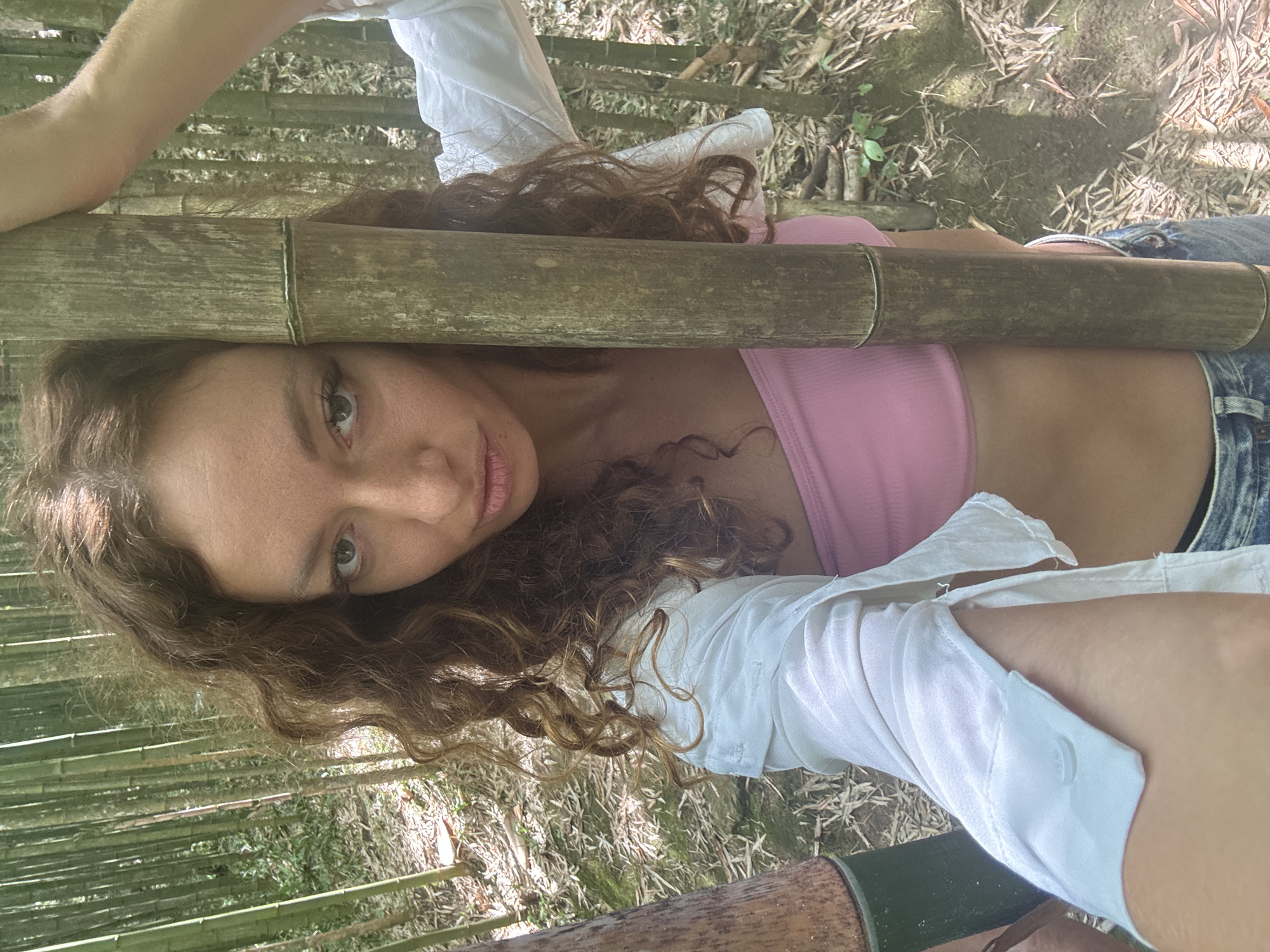 A woman with curly brown hair lies on her back in a bamboo forest, likely located in Ngã Ba Kim, Vietnam. She wears a pink top, denim shorts, and a white shirt, and appears to be resting on bamboo poles. The surrounding environment consists of dense bamboo stalks and ground vegetation.
