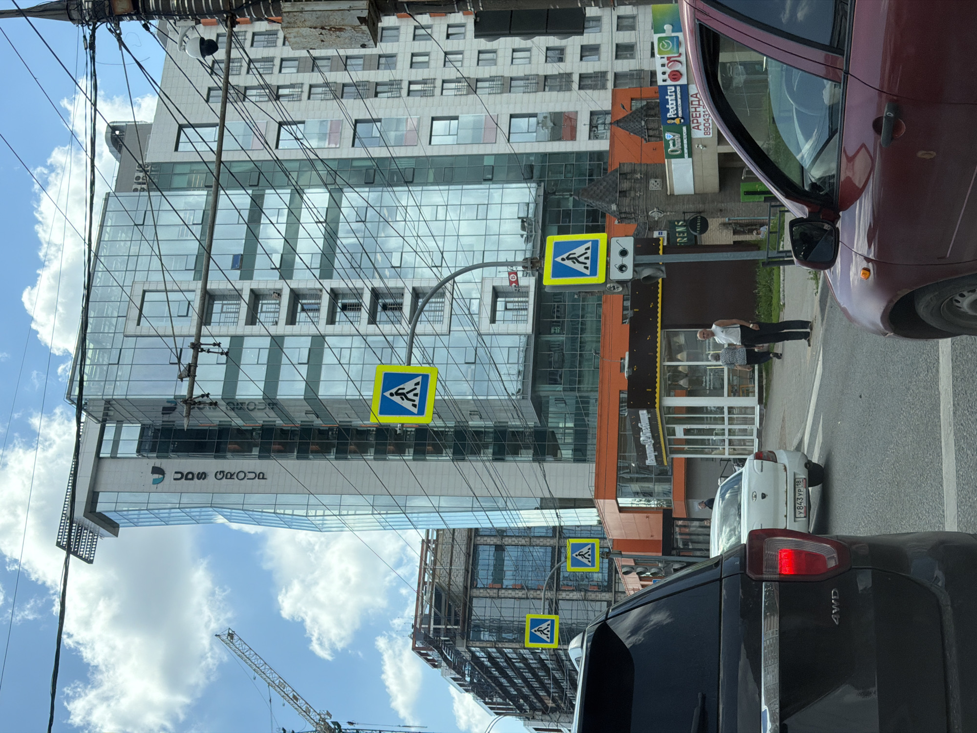 In Izhevsk, Russia, this image captures a street scene dominated by modern architecture and traffic. A tall, multi-story building with a reflective glass facade, bearing the "UDS GROUP" logo, stands prominently. Adjacent to it is another building, partially under construction, with exposed structural elements. Several pedestrian crossing signs are visible, indicating a busy intersection. In the foreground, the rear of a dark SUV and the side of a maroon car are visible. Several individuals are present on the sidewalk, some walking and others standing. The sky is partly cloudy.