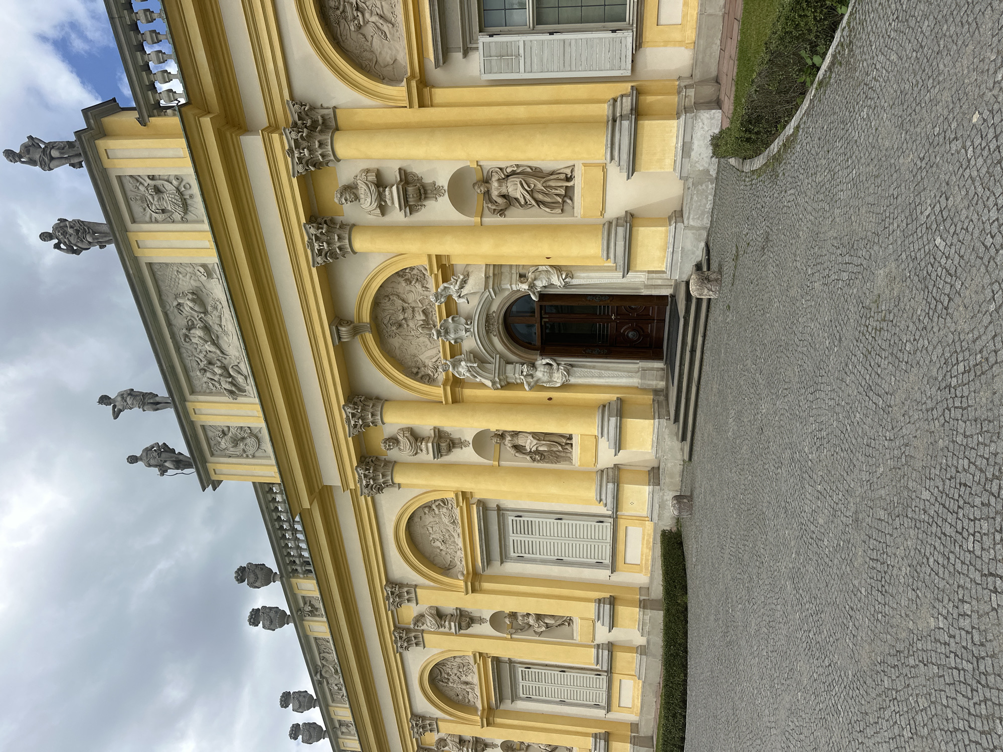 The image shows the facade of the Wilanów Palace in Warsaw, Poland. The palace is painted a pale yellow and features ornate classical architectural elements. Sculptures are placed on the roofline and in niches along the facade. A cobblestone courtyard is in the foreground, with a manicured hedge bordering the right side of the image. The sky is overcast with clouds. There are no people visible in the image.