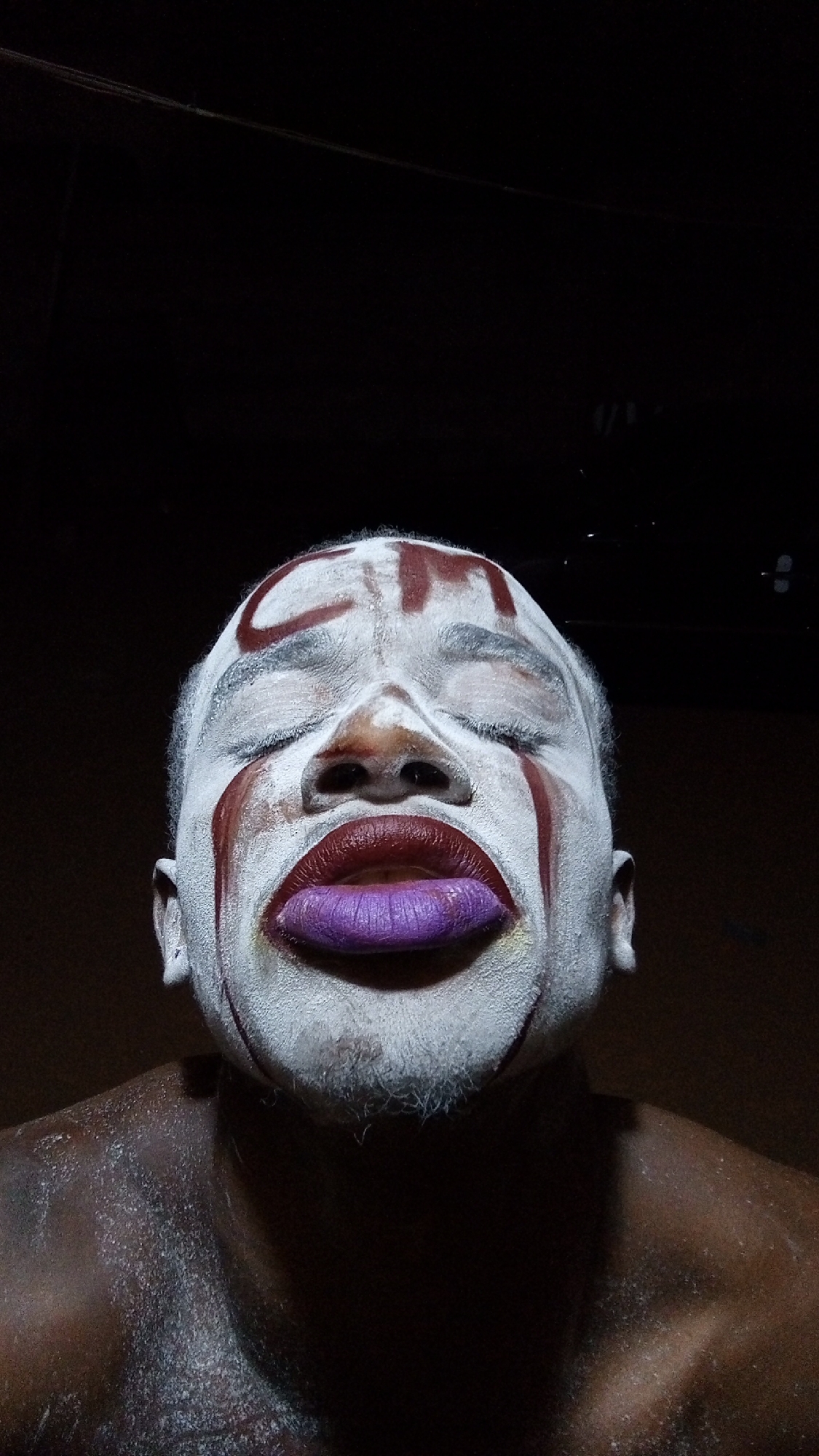The image presents a striking close-up portrait of a person from Onitsha, Nigeria, featuring elaborate face and body paint against a dark background. The individual's face is predominantly covered in white paint, with bold reddish-brown markings forming a symbolic pattern across the forehead. Streaks of the same reddish-brown paint drip from the inner corners of the closed eyes, resembling tears or ritualistic markings. The person's eyes are closed, and the head is tilted slightly upwards, conveying a sense of introspection, reverence, or dramatic expression.
A notable detail is the contrasting lip makeup: the lower lip is a vibrant purple, while the upper lip is a darker maroon or red. The lips are slightly parted, revealing a glimpse of what appears to be the tongue or inside of the mouth, also purple. Light stubble on the chin and jawline is visible beneath the white paint. The upper body, including the shoulders and neck, is also seen, with dark skin dusted with white powder or paint, suggesting a broader application of body art.
The scene is dramatically lit, highlighting the textures of the paint and skin, while the background is completely dark, indicating either a night setting or a controlled, dimly lit indoor environment. The image captures a moment of performance, cultural ritual, or artistic expression. No text is visible in the image.