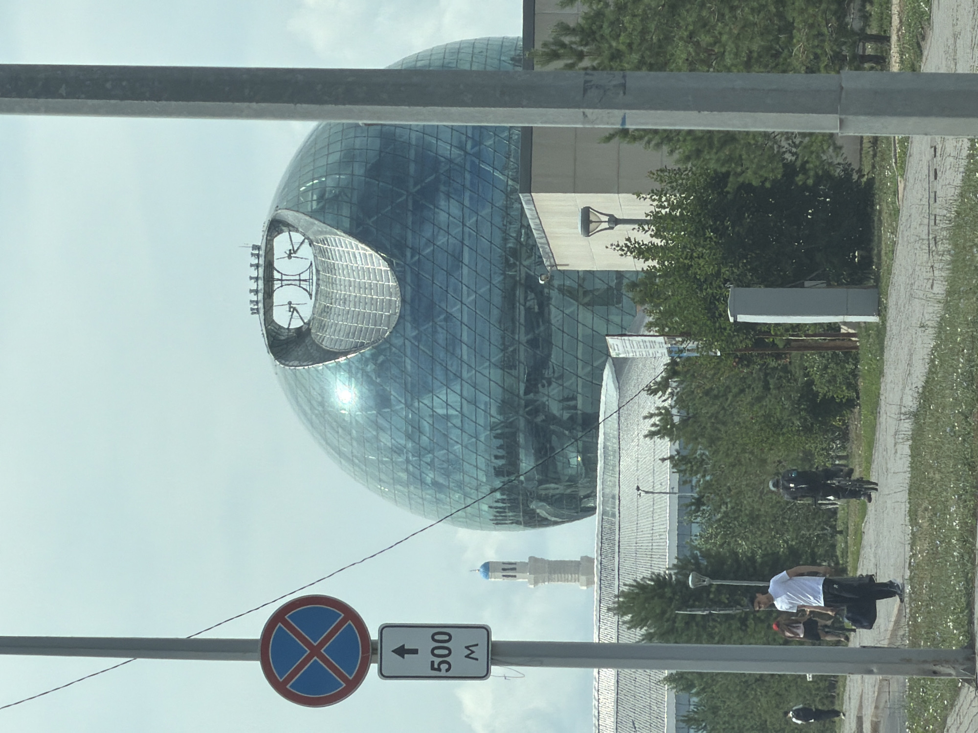 The image captures a view of Astana, Kazakhstan, featuring the prominent Khan Shatyr Entertainment Center, a large, spherical, glass-domed structure. In the foreground, a grey pole stands vertically, accompanied by two traffic signs: a circular "no parking" sign with a blue background and red cross, and a rectangular sign with an upward arrow and the text "500 M". A paved walkway runs horizontally across the mid-ground, where several people are visible. On the left, a person walks away from the camera, carrying a bag. Further down the path, a man in a white t-shirt and dark pants walks towards the camera, also carrying a bag. A person on a bicycle with a backpack rides away from the camera on the right side of the path. In the background, a minaret with a blue dome rises above a low-rise building. Green trees and shrubs are scattered throughout the scene, with the sky appearing overcast and bright.