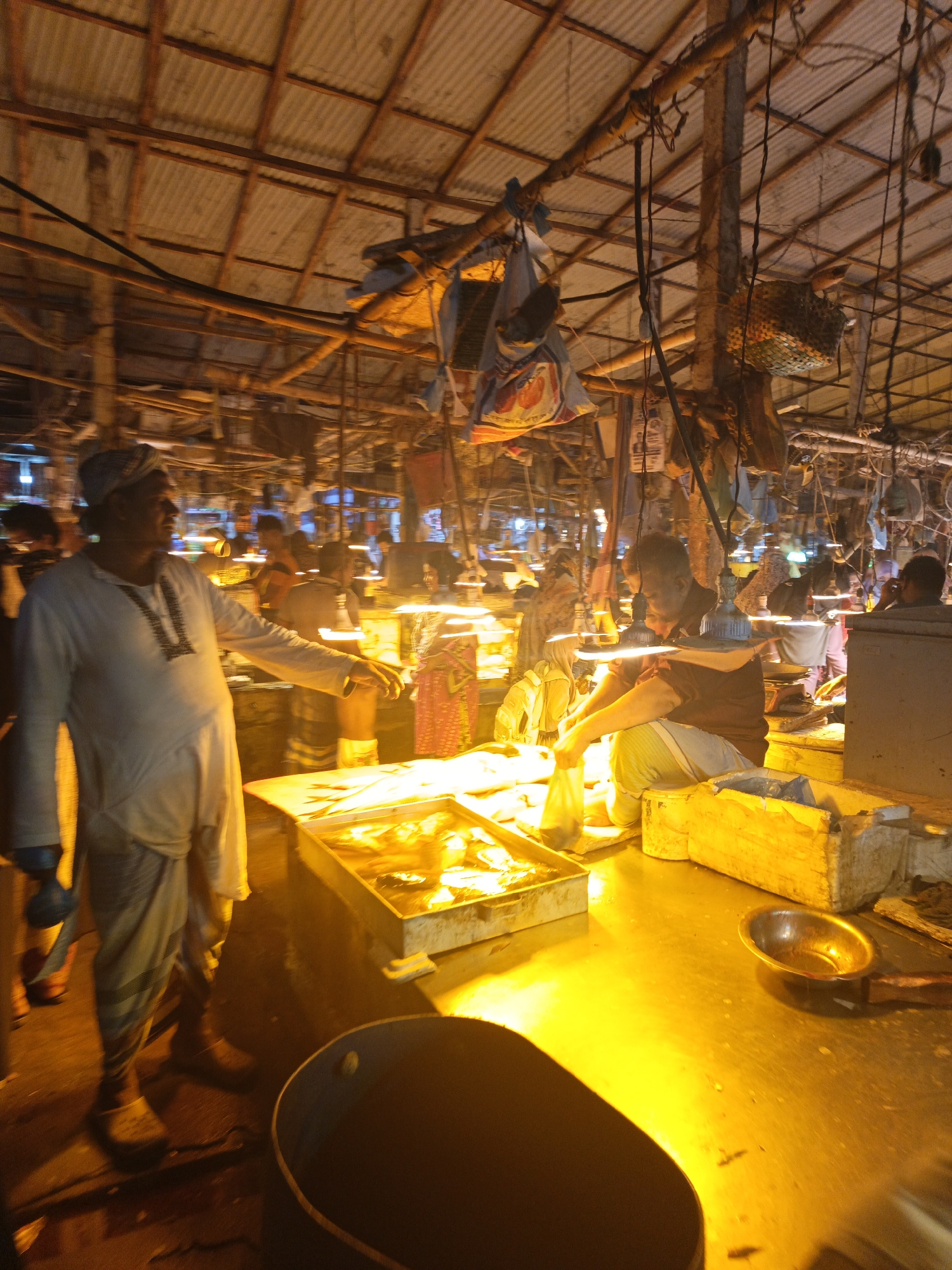 The image captures a bustling fish market in Dhaka, Bangladesh. The scene is illuminated by harsh, warm lighting, casting long shadows and highlighting the activity within the market.
The foreground is dominated by a large, stainless steel counter. A dark, cylindrical container sits on the lower left corner of the frame. A shallow metal pan is present on the right side of the counter. A man with a turban and a light-colored tunic is standing at the counter, gesturing towards a rectangular container filled with fish.
Behind the counter, another man is seated, handling a plastic bag. In the background, numerous other individuals are visible amidst a maze of tables, overhead lights, and various hanging objects, possibly bags or baskets.
The market's infrastructure is characterized by a corrugated metal roof supported by a network of wooden beams and poles. Exposed wiring and hanging light fixtures contribute to the raw, utilitarian atmosphere of the market.
