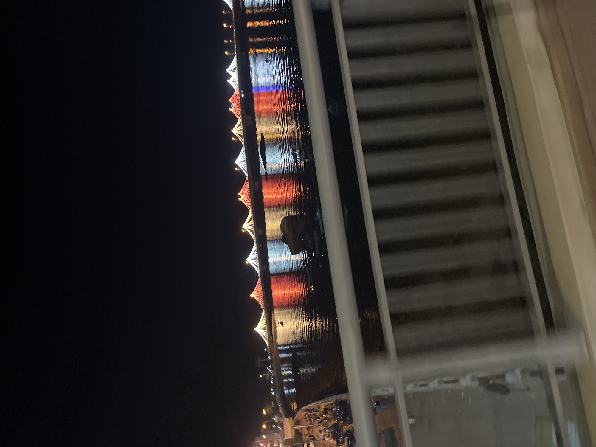 A night scene from Bac Nha Trang Ward, Vietnam, viewed through a window with closed blinds. Outside the window, a body of water reflects strings of lights hung along the shore. The lights are in a repeating pattern of white and red. Buildings and vehicles are visible near the base of the string lights.