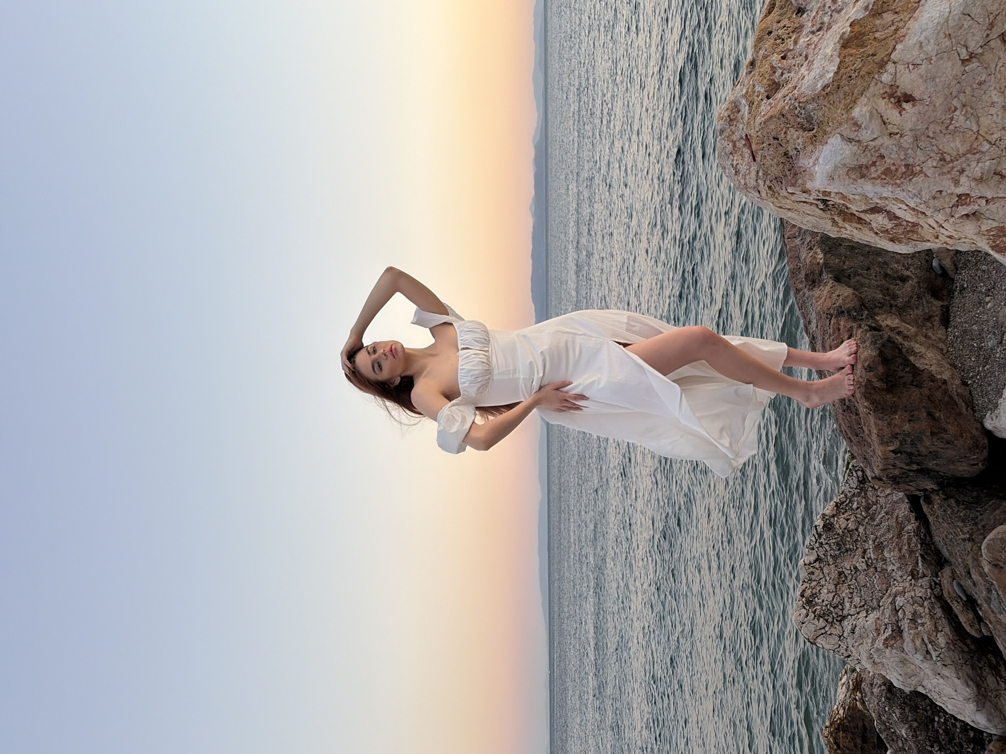 The media file displays a woman in a white dress lying on her side on rocks by the sea at Paleo Faliro, Greece. The woman is positioned with her head on her hand, looking towards the viewer. Her body is draped in a flowing white dress, and her feet are visible at the edge of the rocks. The background features the sea with gentle waves and a distant horizon line where the sky transitions from a pale blue to a soft peach hue, indicating it is either dawn or dusk. The setting includes rough, textured rocks in the foreground and mid-ground, with the sea extending into the background. The overall mood is serene and peaceful, with the natural beauty of the location and the gentle lighting contributing to a tranquil atmosphere. There is no visible text in the media file.