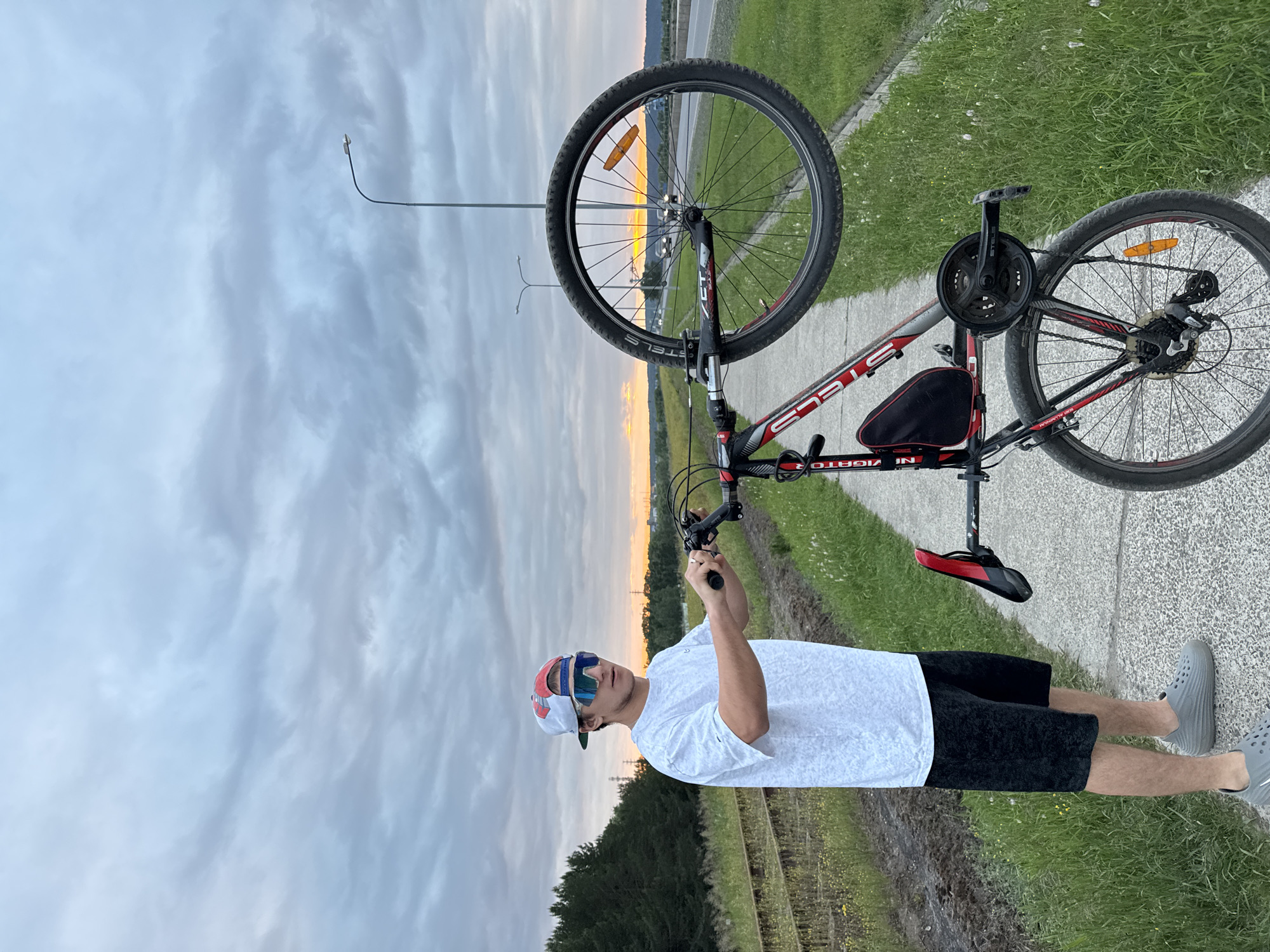 In Chaikovsky, Russia, a young man stands on a paved path next to an abandoned railroad track. He is wearing a white t-shirt, black shorts, a baseball cap, and sunglasses. He holds a bicycle above his head with the front wheel extended upwards. The bicycle is red and black, with a black saddle bag attached to the frame. The background shows a cloudy sky, a highway with cars, and a forest. The scene appears to be at dusk, with the sun setting on the horizon.