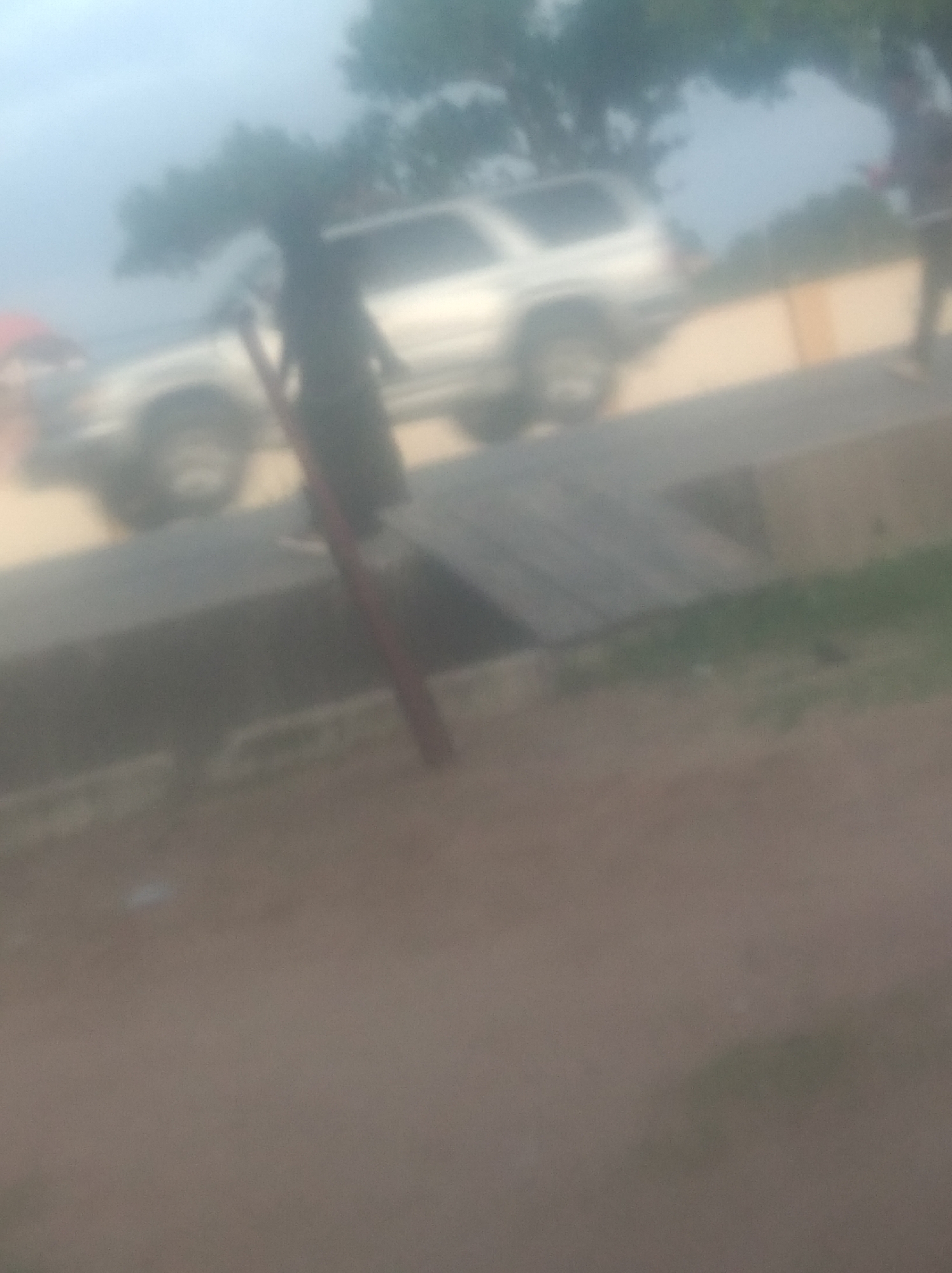 The image is a blurry, overexposed photograph of an outdoor scene in Jalingo, Nigeria. In the background, a silver SUV is parked on a dirt road. A person, possibly a man, is standing beside the vehicle, partially obscured by a tree. Another person, wearing a red shirt, is visible to the left of the SUV. The ground is covered in a mixture of dirt and gravel, with some patches of green vegetation. The sky is overcast and hazy, suggesting it may be a cloudy or dusty day. The overall impression is of a casual gathering or a brief stop on a journey, set against the backdrop of a typical Nigerian roadside environment.