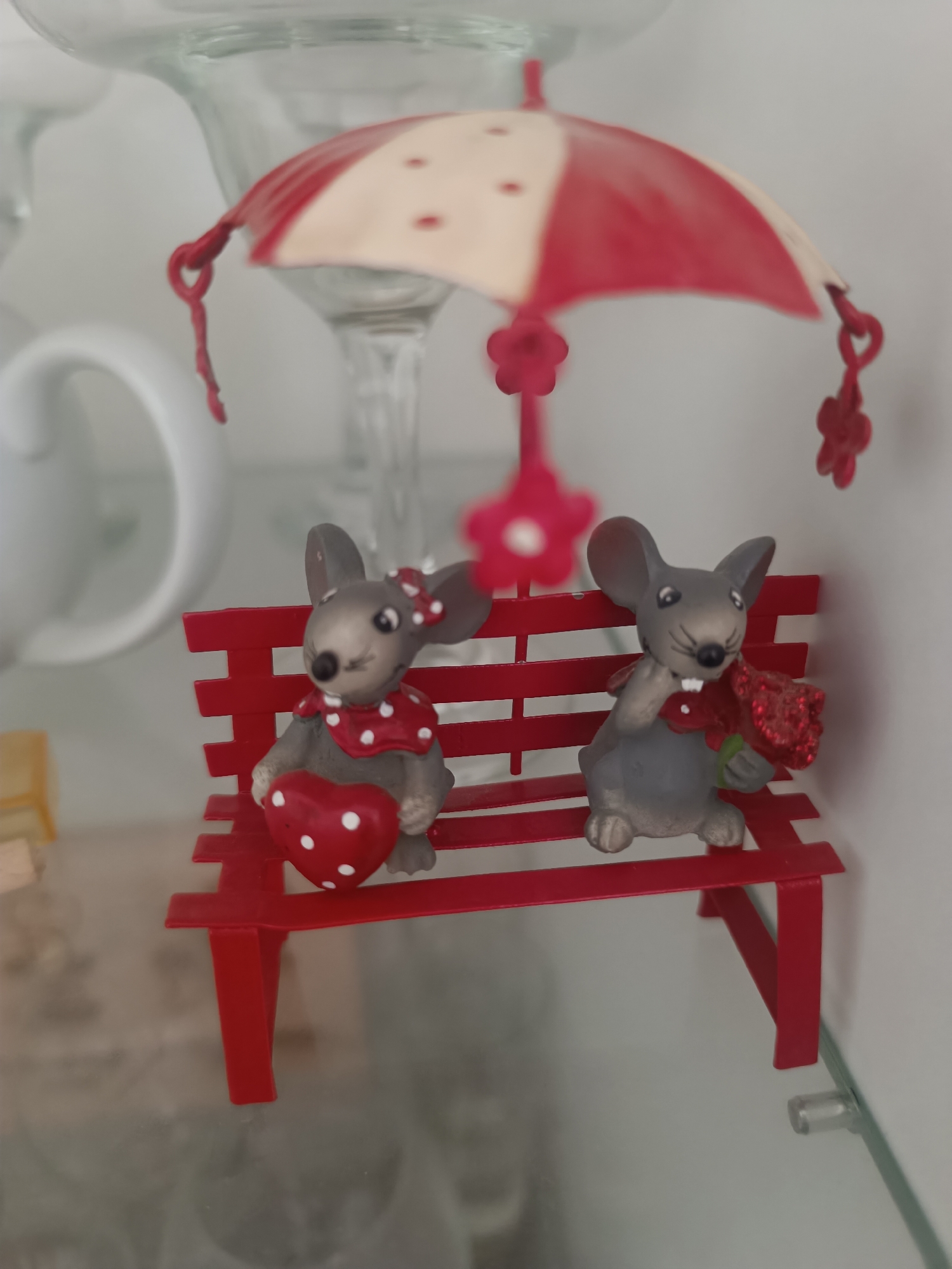 A close-up image displays two decorative mouse figurines seated on a miniature red slatted bench. Above them, a small red and off-white striped umbrella figurine hangs, suspended by a red string with flower-shaped embellishments.
The mouse on the left is grey, wearing a red bandana with white polka dots and a red bow on its head. It holds a red heart figurine with white polka dots. The mouse on the right is also grey, wearing a red collar, and holds a small bouquet of red, glittery roses with green leaves. Both mice have black eyes and prominent white teeth.
The red bench is constructed with a slatted back and seat, and four legs. It rests on a clear glass shelf, which appears to be part of a display cabinet. In the background, out of focus, are various household items, including clear glass stems of what appear to be glassware (possibly wine glasses or similar vessels) and a white ceramic handle from a mug or cup. The lighting suggests an indoor setting. The specific location within Varna, Bulgaria, is not discernible from the visual information provided in the image.