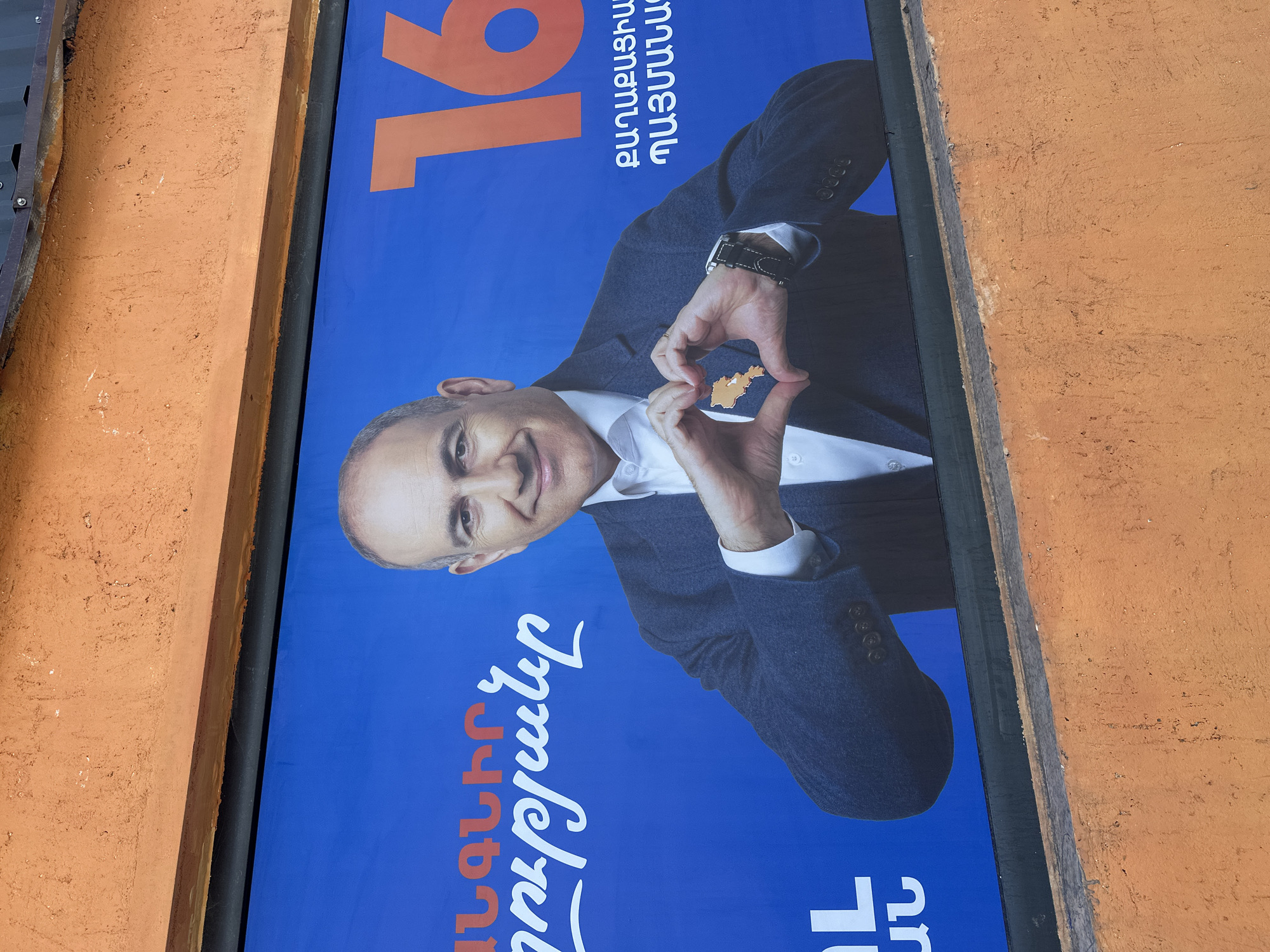 The image captures a political campaign poster prominently displayed in Yerevan, Armenia. The poster features a portrait of a bald man, possibly a politician or public figure, who is smiling and making a heart shape with his hands. Inside the heart, there is a smaller shape that resembles a map. He wears a dark suit, a white shirt, and a wristwatch. The poster has a bright blue background with white and orange Armenian text. A large number '16' is displayed in orange. The poster appears to be mounted on an exterior wall, situated between a black metal frame and an orange-colored wall with a rough, textured surface.