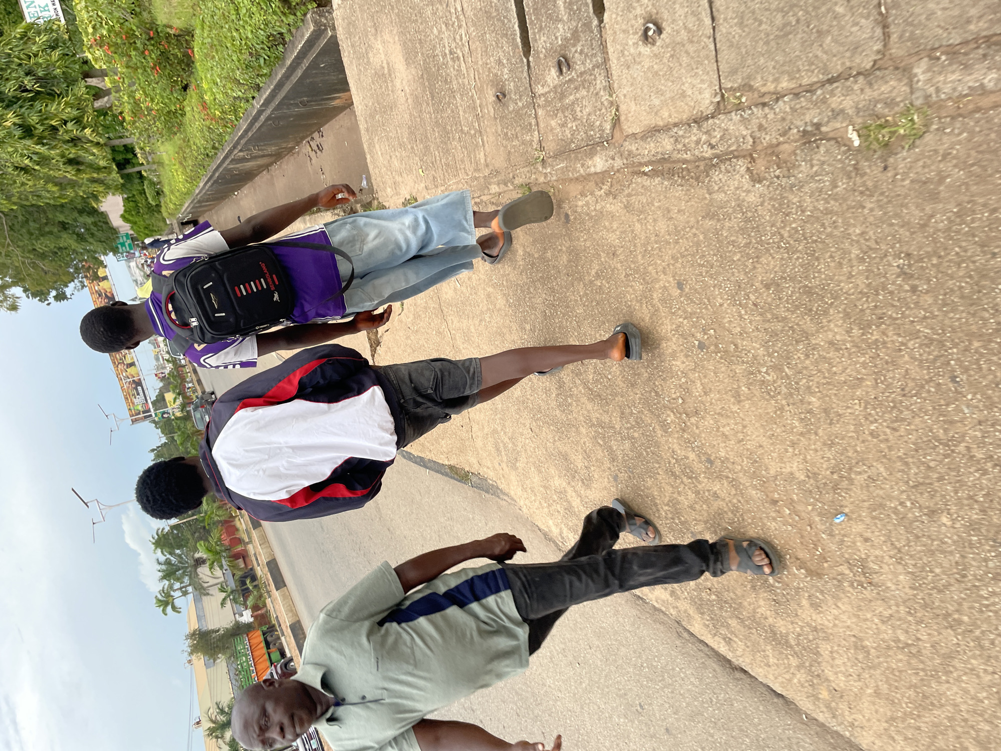 In Jos, Nigeria, on a cloudy day, three individuals are walking along a paved sidewalk adjacent to a road. The person in the foreground, an adult male with a light skin tone, is wearing a short-sleeved, light green polo shirt with a dark blue stripe on the left side, black pants, and dark sandals. He is walking away from the camera. Behind him, two younger individuals, both appearing to be male, are also walking in the same direction. The person in the middle is wearing a jacket with white, red, and black panels and dark shorts. The person on the right has dark skin and is wearing a purple and white patterned t-shirt, light blue wide-leg pants, and dark sandals with an orange accent on one of the straps. He is carrying a black backpack with red detailing. The sidewalk is made of concrete and shows some signs of wear, with drainage grates visible. The road next to the sidewalk has visible tire marks and markings. In the background, there are buildings, palm trees, lush green vegetation, and streetlights. A billboard advertising "ENI CON... PARK" is partially visible on the right. A green truck is also visible on the road. The overall atmosphere is that of a typical urban environment in Nigeria.