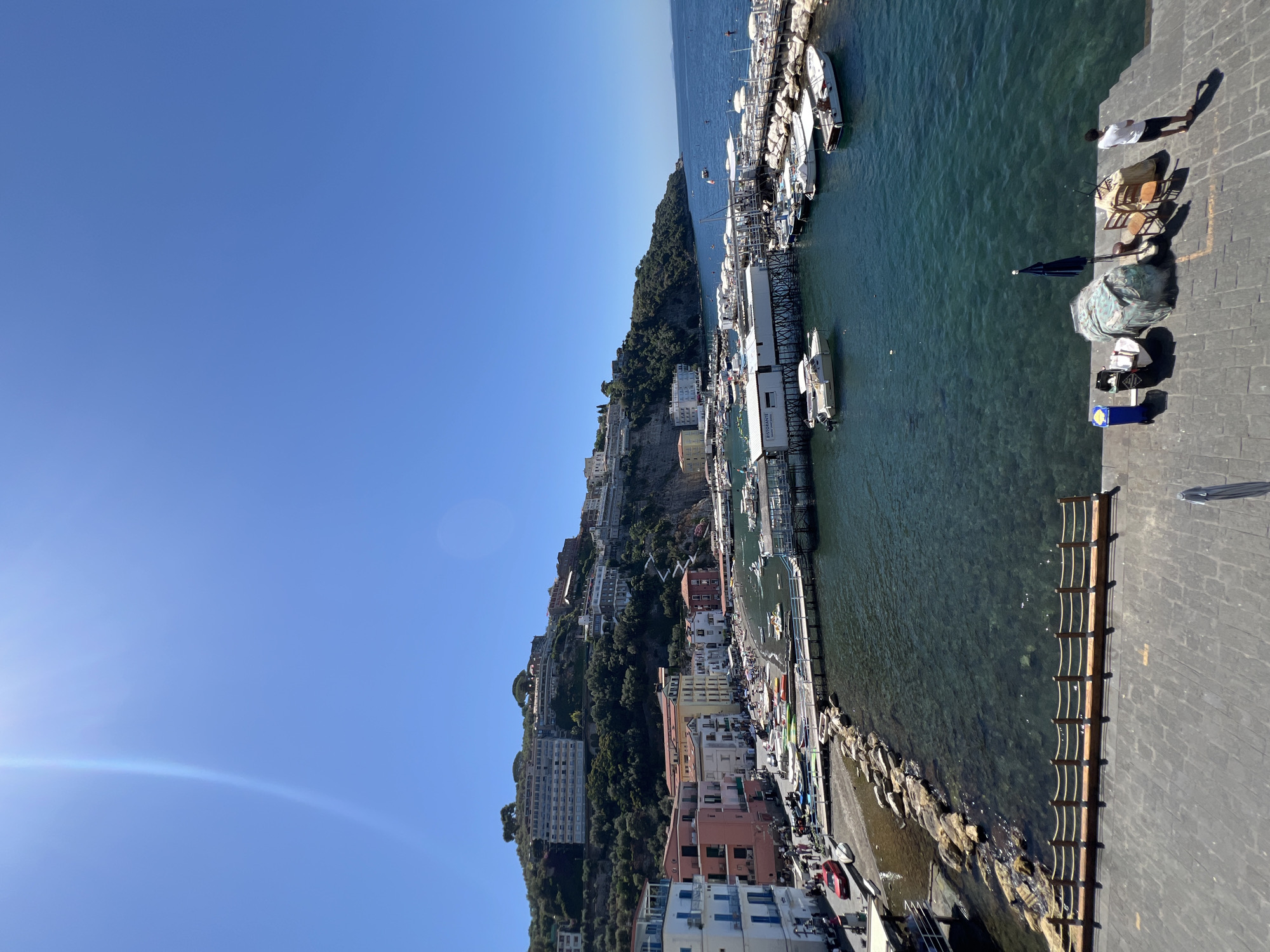 The image shows a scenic view of a bay in Sorrento, Italy, on a clear, sunny day. The foreground is a paved walkway with a wooden rope fence. On the right side of the walkway, there are several objects including a blue trash bin, a black trash bin, a covered object, a stool, two chairs, and a person standing.
The bay itself is filled with clear, dark blue water, populated by numerous boats of various sizes. Some are docked at piers, while others are anchored. Along the edge of the bay, there is a small beach with people relaxing and several colorful kayaks and paddleboards.
Behind the beach, a row of buildings lines the waterfront. These buildings are colorful, with pastel facades and balconies. Further up the hillside, more buildings and hotels are visible, nestled among lush green trees. The hillside slopes steeply towards the sea, creating a dramatic backdrop. The sky is a vibrant, cloudless blue, with the sun casting a bright light, creating lens flares. The overall scene depicts a picturesque coastal town with a bustling harbor and a lively beach.