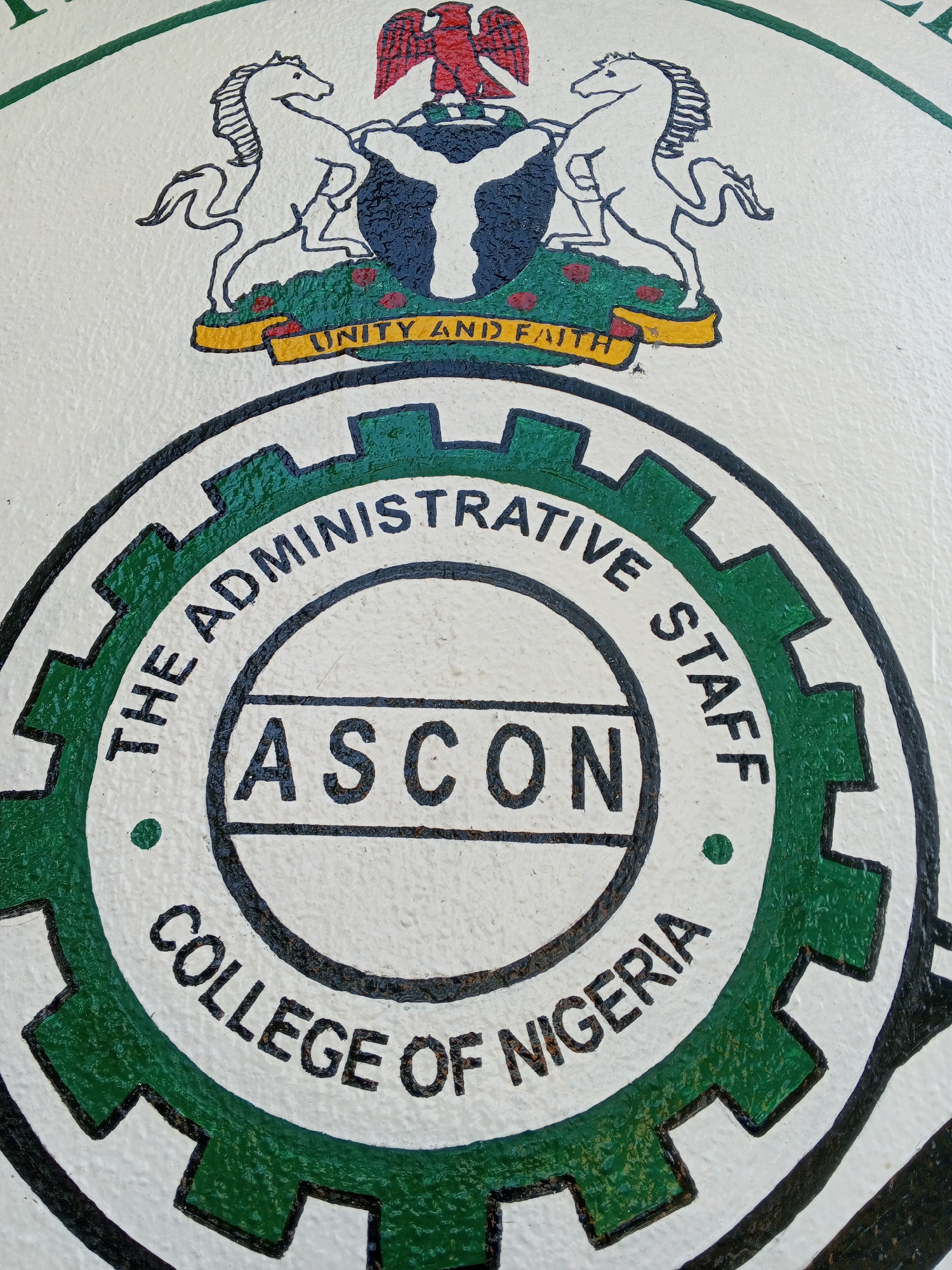 The image shows a painted emblem. At the top is the coat of arms of Nigeria. Below the coat of arms is a circular seal with a gear-like border. In the center of the seal, the acronym "ASCON" is printed between two horizontal lines. Encircling the center is the text "THE ADMINISTRATIVE STAFF COLLEGE OF NIGERIA". It is likely that this emblem is located in Tawpo, Nigeria, given the presence of "COLLEGE OF NIGERIA". The scene depicts a symbol representing the Administrative Staff College of Nigeria.