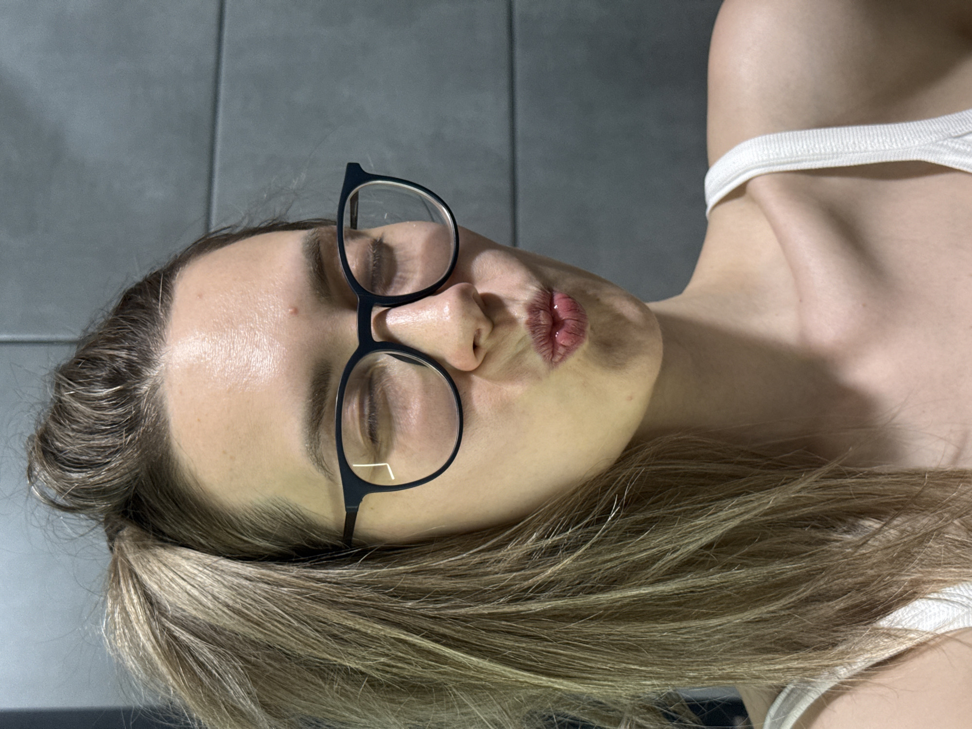 The image is a close-up selfie of a woman with light blonde hair. She is wearing black-framed glasses and a white tank top. Her eyes are closed, and she is making a kissing face. The background is a gray tiled wall. There is no evidence to suggest that the location of the image is in the city of Chaikovsky, Russia.