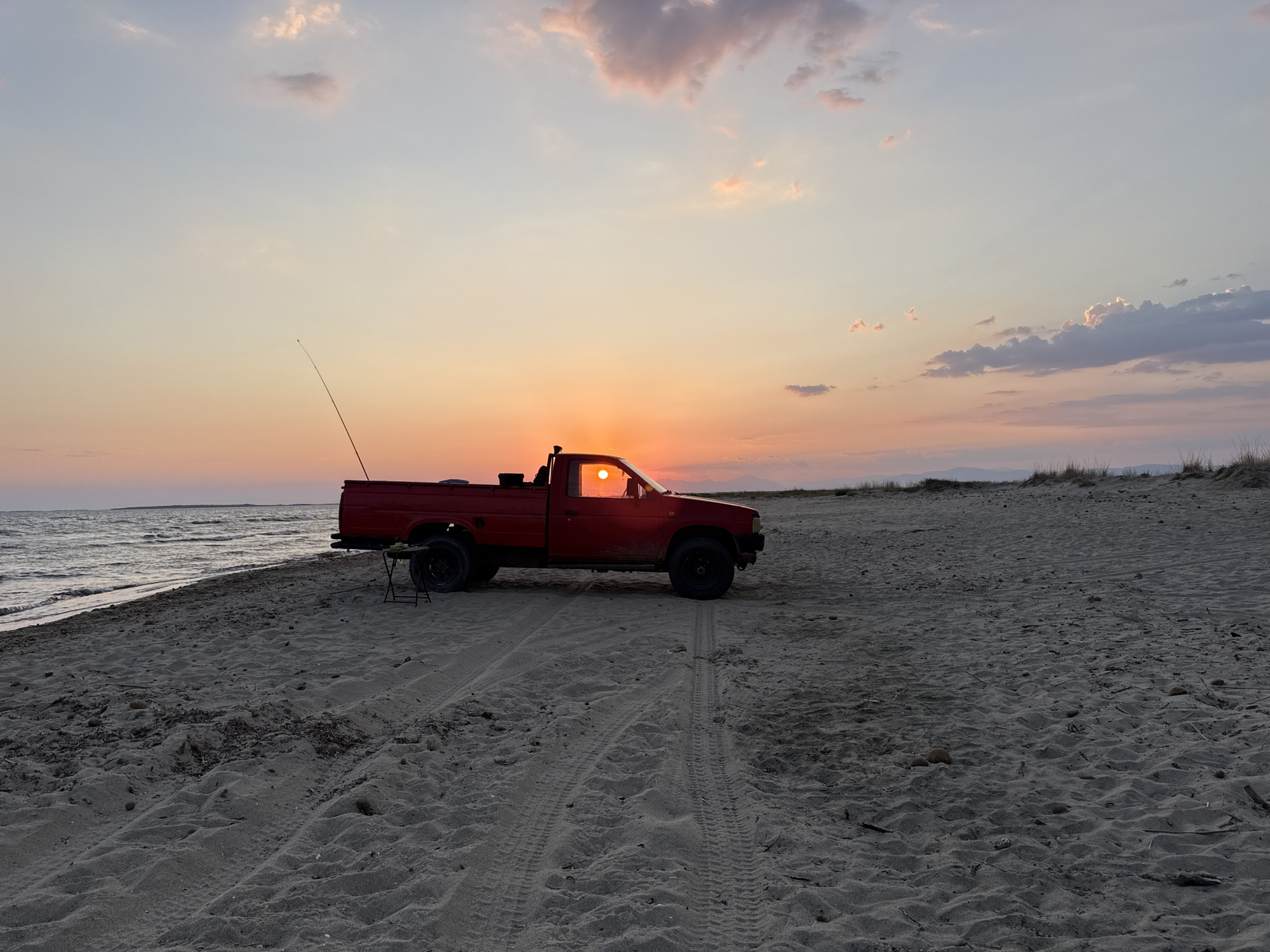 In Imeros, Greece, a red pickup truck is parked on a sandy beach near the sea during sunset. The sun, a bright orange orb, is partially visible through the truck's driver-side window, casting a warm glow on the scene. The sky is a gradient of soft blues, oranges, and pinks, with scattered clouds. Tire tracks are visible in the sand behind the truck. A small folding table and chair are set up next to the truck. The ocean waves gently lap at the shore. In the distance, a faint line of hills is visible. The overall atmosphere is tranquil and serene.
